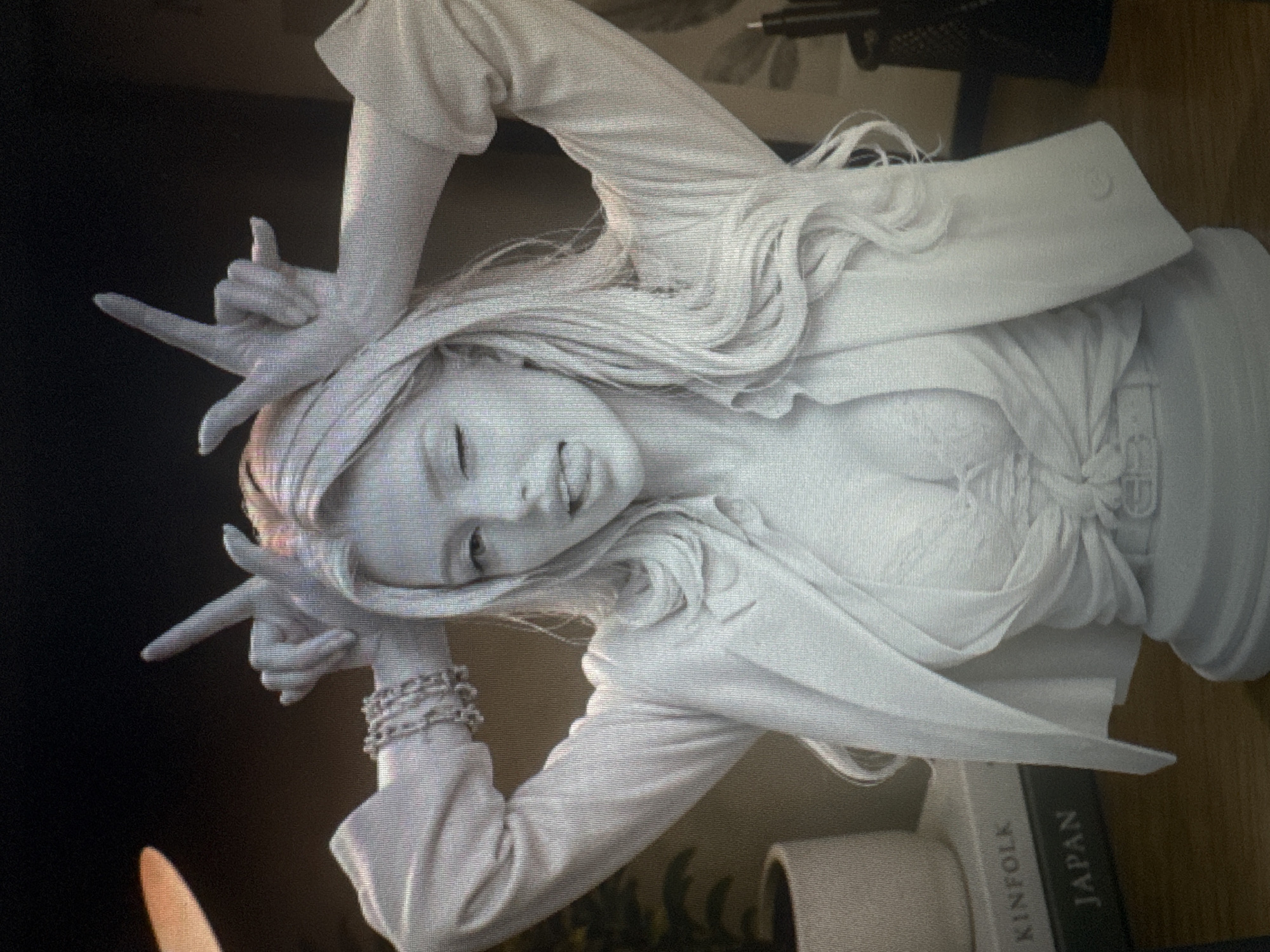 The media file shows a close-up, angled shot of a white bust sculpture of a young woman with long, flowing hair. The sculpture is a pale, matte white and appears to be made of plaster or a similar material. The woman has her head tilted back slightly, with her eyes closed and a slight smile on her face, suggesting a relaxed or playful mood. Her hair is depicted as very long and wavy, falling around her shoulders and over her arms.
She is wearing a jacket that is open, revealing a draped top underneath. Her arms are raised, with her hands positioned in a gesture that resembles the "rock on" or devil horns sign, with two fingers extended upwards and the thumb and other fingers curled in. She has a bracelet on her left wrist, which appears to be made of small, interconnected metallic elements.
The background is dark and out of focus, with some hints of a wooden surface and possibly other objects, including a book that has text visible. The visible text on the book reads "KINFOLK" and "JAPAN." The lighting in the scene creates subtle shadows that highlight the textures and contours of the sculpture. The overall impression is of an artistic representation of a youthful and energetic individual.