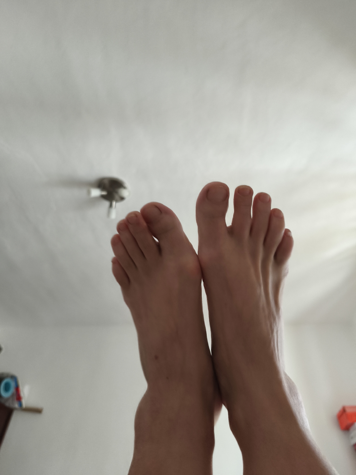 The image shows a close-up, low-angle view of a person's bare feet and lower legs against a white ceiling with a light fixture. The feet are positioned with the toes pointing upwards, and the soles of the feet are not visible. The skin appears fair, and there are no visible blemishes or adornments such as nail polish or jewelry. The ceiling is textured and shows subtle shadows, suggesting ambient light. In the background, out of focus, is a wall that appears to be white or a very light color. On the left side of the background, a blurry shelf with some objects, possibly toys or storage containers, can be seen. On the right side of the background, a blurred red and white object is visible, which might be a box or container. The overall impression is of a domestic indoor setting. The lighting suggests it could be daytime, but there are no strong indications of the time of day. There is no visible text in the image. The activity appears to be resting or posing with feet elevated.