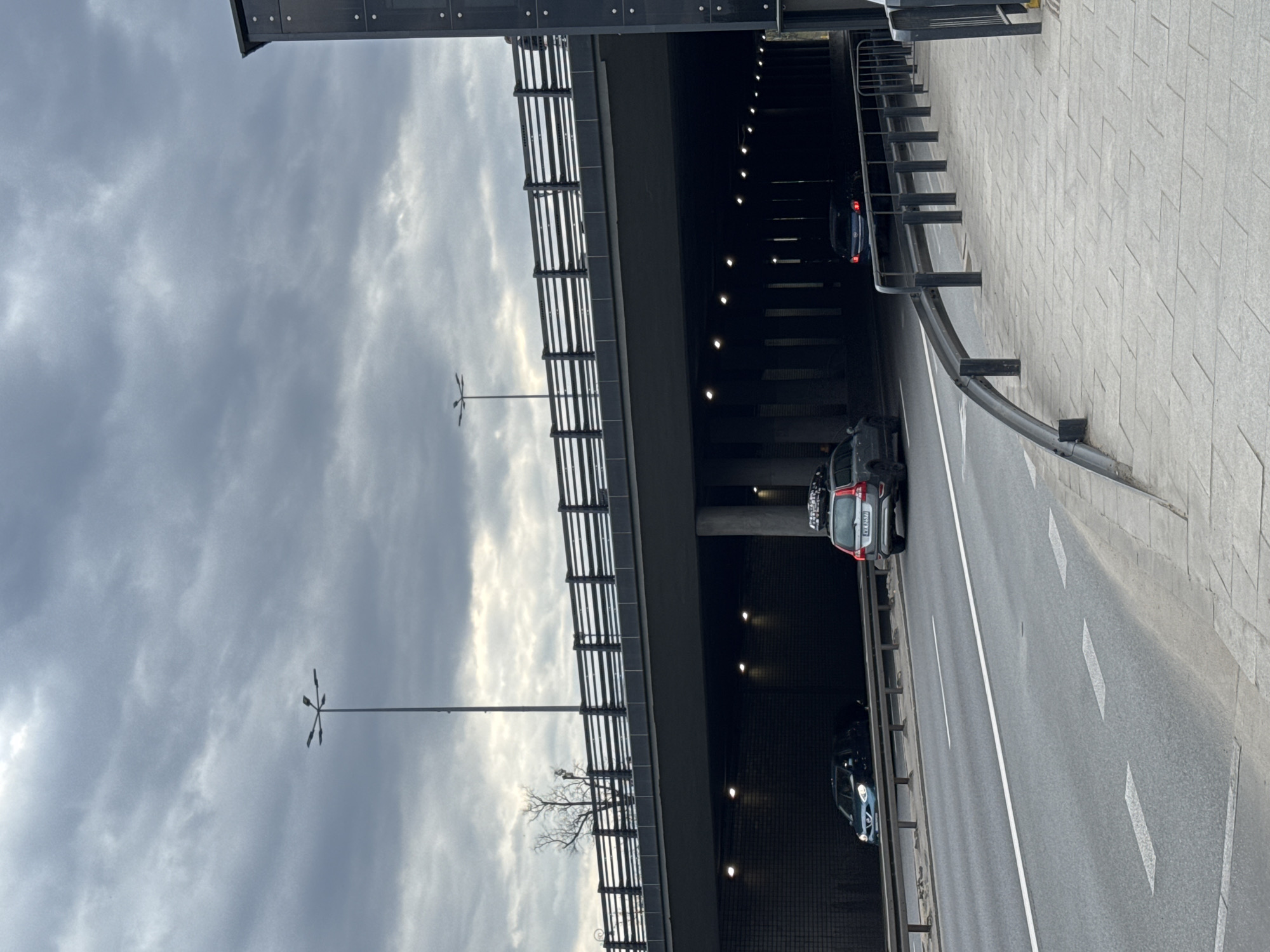 An eye-level, color, outdoor shot depicts the Powiśle Viaduct in Warsaw, Poland. The viaduct has two overpasses and runs over a gray asphalt road with a solid white center line. There are three vehicles in view. The viaduct structure appears to be constructed of dark gray concrete and metal railings. On the right side of the road is a light gray concrete sidewalk. Light poles with abstract geometric configurations are visible on the left. The sky is a uniform dull gray.