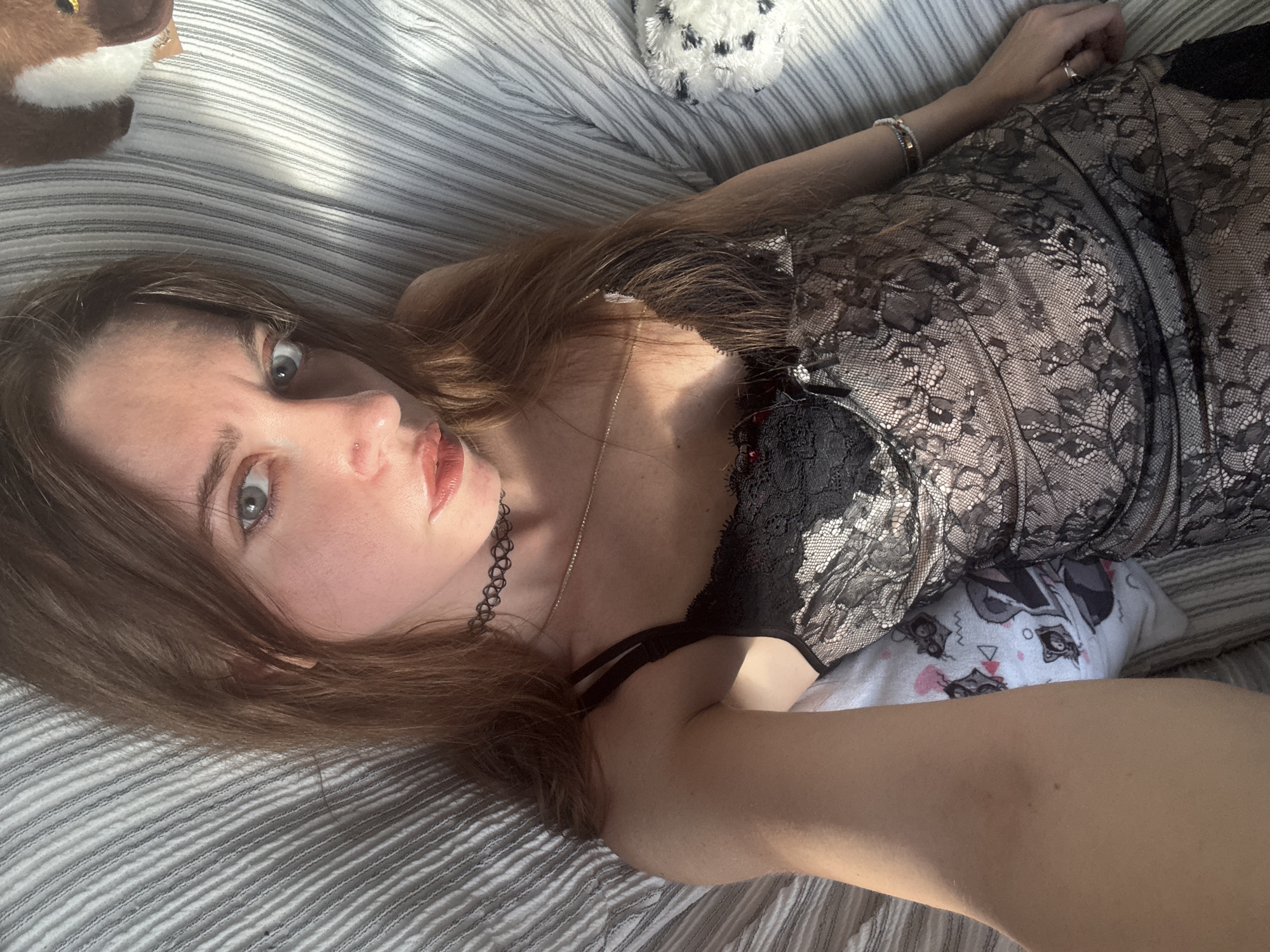 A young woman with light brown hair is lying on a striped surface, possibly a bed or couch. She is looking directly at the camera with a neutral expression. She is wearing a black lace dress with thin straps and a choker necklace. The lighting suggests it is daytime, with sunlight casting shadows on her. The background is blurred, but a stuffed animal can be seen to the right. The image appears to be a selfie taken from a slightly elevated angle.