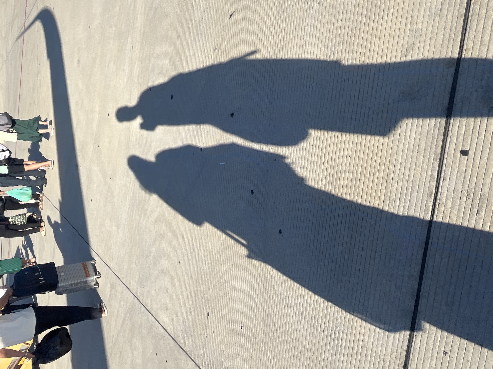 The image shows a sun-drenched concrete tarmac, likely at Cam Ranh International Airport in Cam Ranh City, Vietnam, given the context. Two long, stark shadows of people are cast across the ribbed surface of the tarmac. The shadow on the left appears to belong to a person holding a bag, while the shadow on the right is of a person with what seems to be a phone or camera raised. In the background, the lower legs and feet of several individuals are visible, some carrying luggage, suggesting they are travelers. The bright sunlight indicates it is daytime.