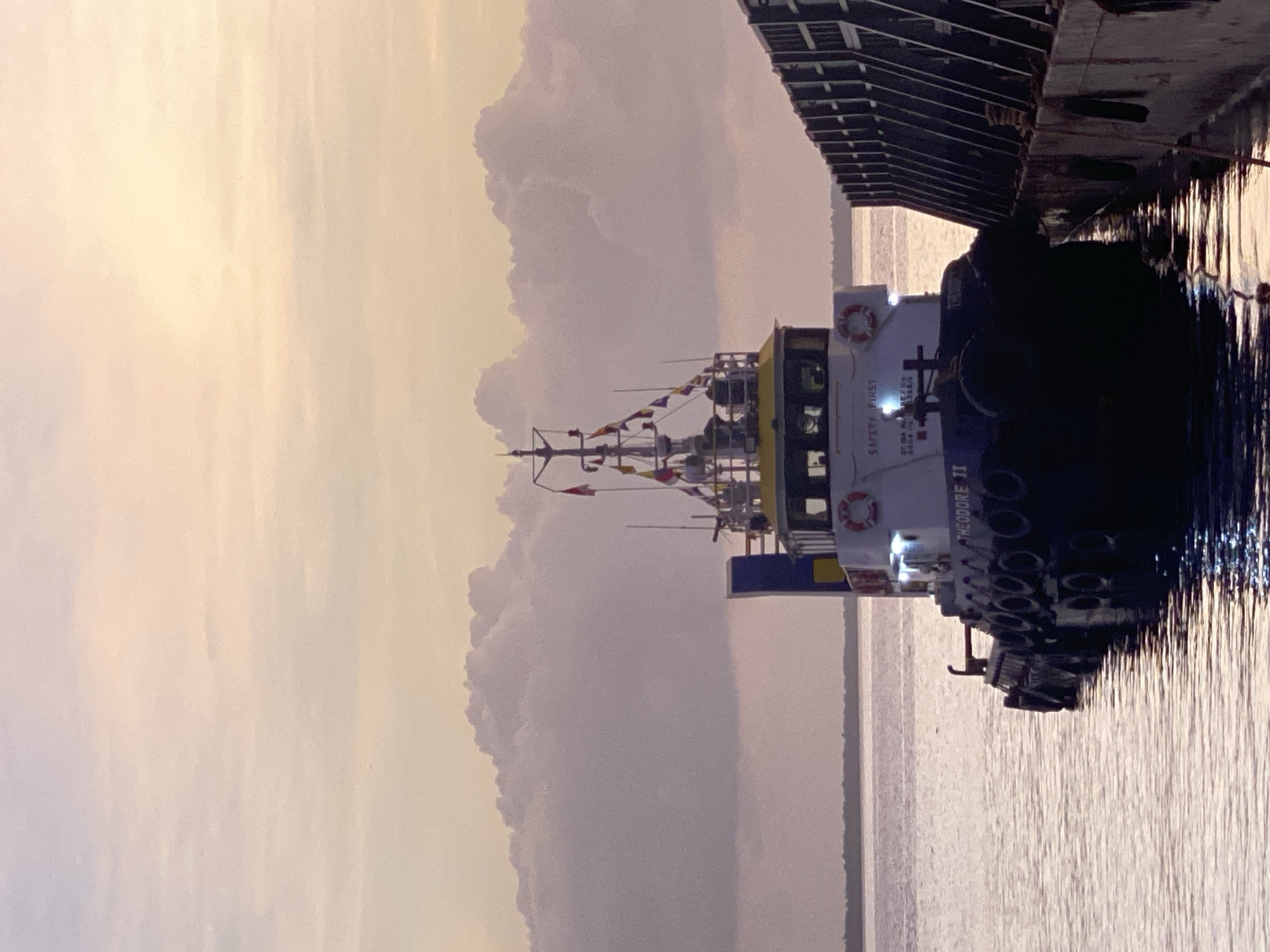 A tugboat named "THEODORE II" is docked next to a barge at sunset in the waters of Indonesia. The sky is filled with dramatic, illuminated clouds, casting a soft orange and pink hue over the scene. The tugboat has a white cabin with "SAFETY FIRST" visible on it, and a red life preserver. Several flags are flying from the tugboat's mast. The barge is dark and appears to be made of concrete. The water is calm, reflecting the colors of the sky. The overall atmosphere is serene and peaceful.