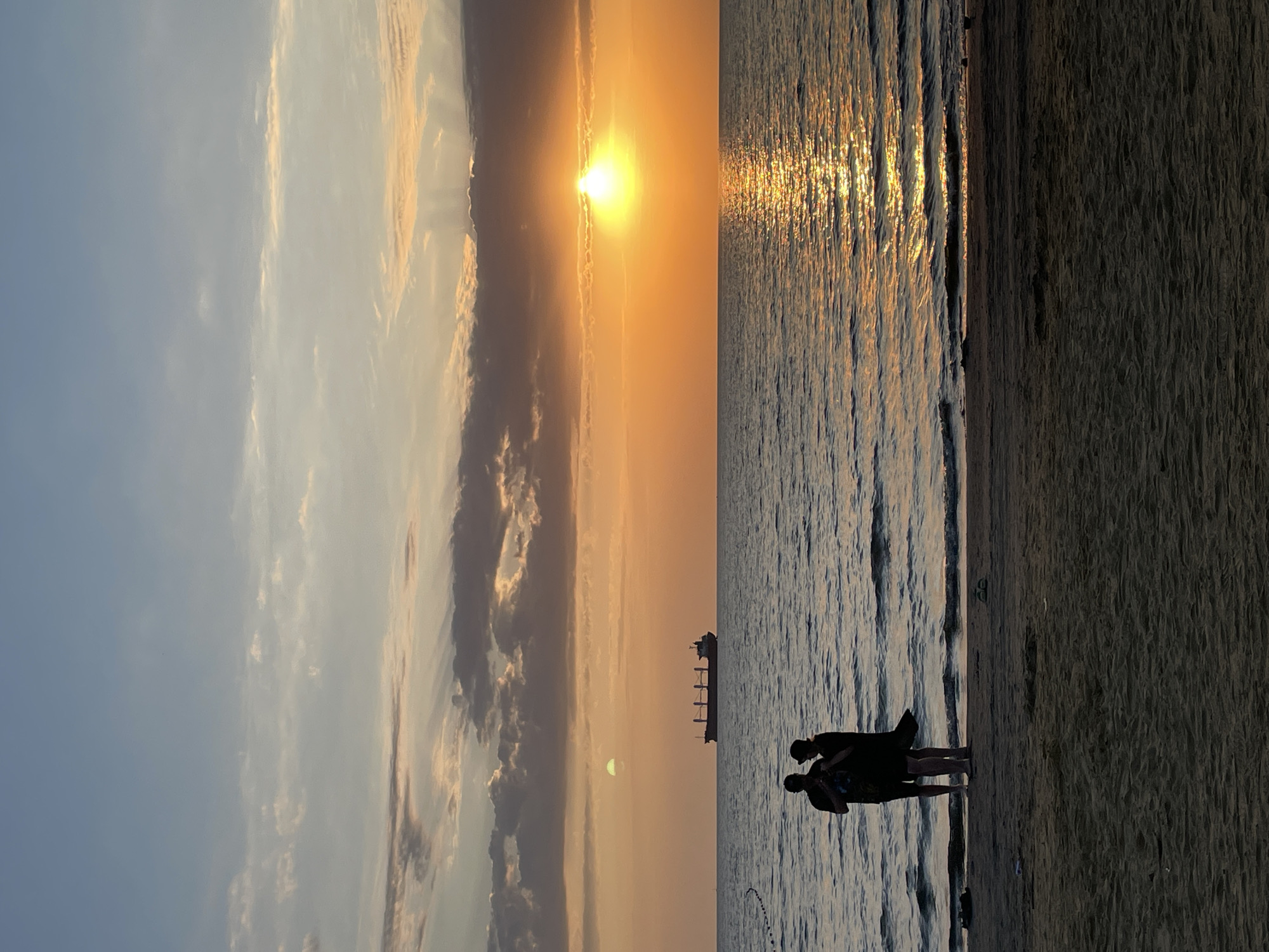 The image captures a serene sunset scene on a sandy beach in Anapa, Russia. Two individuals, silhouetted against the vibrant sky, stand near the water's edge. They are facing the sea, with one person appearing to hold something. A large cargo ship is visible on the horizon, sailing across the calm water. The sun, a bright orange orb, is setting, casting a golden reflection on the rippling surface of the sea. The sky is a gradient of blues and oranges, with scattered clouds adding texture to the celestial canvas. The sand in the foreground is textured, with subtle shadows indicating the time of day. The overall atmosphere is peaceful and contemplative.