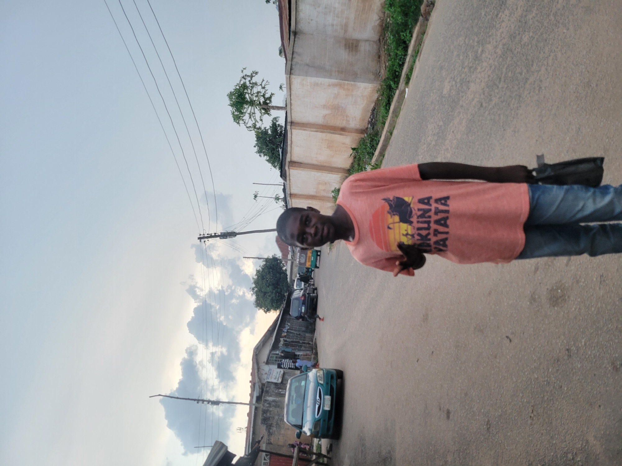 A young person is standing on a paved road, giving a peace sign. The person is wearing a salmon-colored t-shirt with "Hakuna Matata" printed on it and blue jeans, and is holding a black bag. The background shows a street scene with cars, buildings, and power lines. The sky is overcast.
The location appears to be a street in Jos, Nigeria. Evidence includes the style of architecture, the types of vehicles, the style of clothing, and general environment.