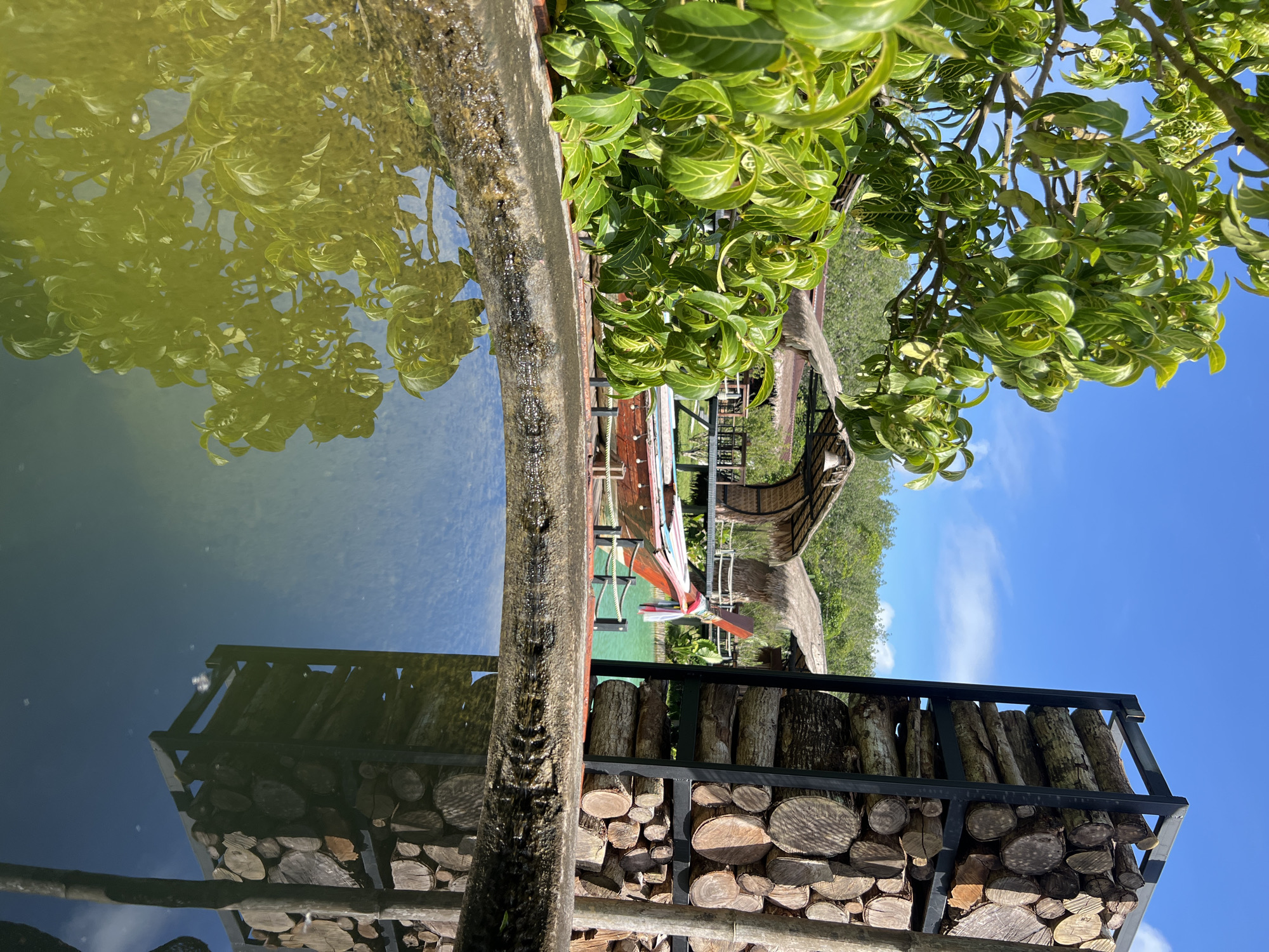 The image depicts a peaceful outdoor scene in a tropical setting, likely in Thailand given the location context. The foreground features a textured, rough-hewn wooden beam, possibly part of a railing or structure, positioned horizontally. Adjacent to it is a body of calm water, reflecting the surrounding greenery and the blue sky. The water has a slight yellowish tint, suggesting it might be a pond or a shallow pool with algae or sediment.
Behind the water, a collection of neatly stacked logs is visible, contained within a black metal frame. These logs vary in size and diameter, showcasing different wood grains and textures.
Further into the scene, a colorful boat with ornate decorations and colorful ropes is partially submerged or docked in the water. The boat has a traditional design, suggesting it could be a longtail boat common in Southeast Asia. Behind the boat, a thatched-roof structure, possibly a hut or a pavilion, is visible amidst lush green foliage. The trees are dense and verdant, typical of a tropical environment.
The sky is a clear, bright blue with a few wispy white clouds, indicating fair weather and likely daytime, possibly mid-morning or afternoon. The overall atmosphere is serene and tranquil, evoking a sense of relaxation and natural beauty. No people are visible in the image, and there is no visible text.