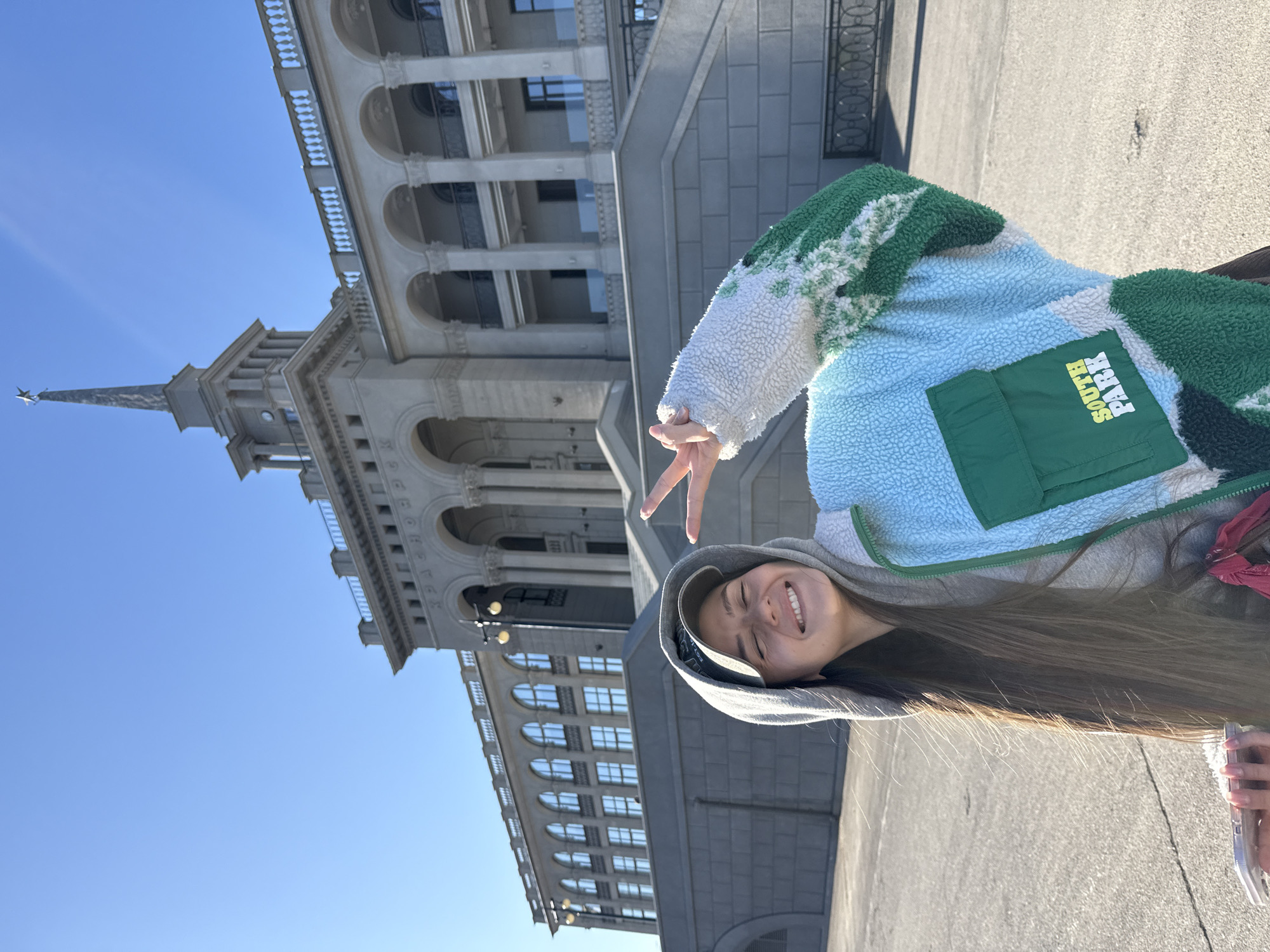 A young woman is posing for a picture in Krasnoyarsk, Russia. She is wearing a gray hooded sweatshirt under a colorful South Park-themed jacket, making a peace sign with her fingers. The building in the background is likely the Yenisey Krasnoyarsk hotel. The sky is clear and blue. The woman is smiling.