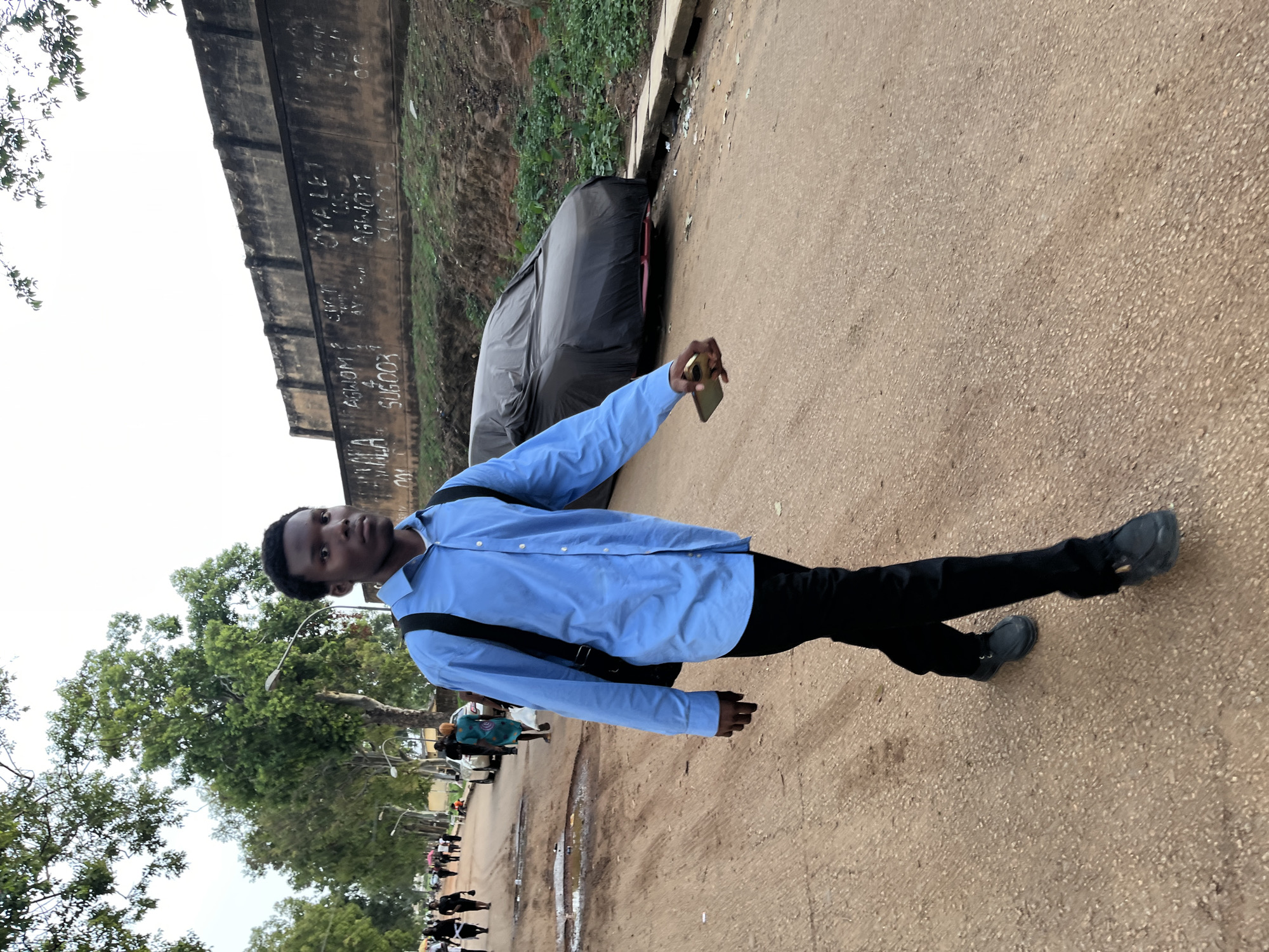 This image captures a young man standing on a dirt-and-gravel road in what appears to be a semi-urban or rural setting in Rafin Par, Nigeria. The man, the central subject, is fair-skinned with short dark hair, wearing a light blue long-sleeved button-up shirt, black trousers, and black shoes. He has a black shoulder bag slung across his body and is holding a gold-colored smartphone in his right hand, extended forward, possibly posing for a photo or taking a selfie/video. His expression is neutral as he looks directly at the camera.
To his right, behind him, is a dark-colored car covered with a black tarp, parked off the road. Further right and behind the car is a low dirt embankment covered with sparse vegetation. Running parallel to the road on the left side, behind the man, is a tall, aged concrete wall covered in white graffiti. Visible text on the wall includes "ABHOM," "SUGOOX," "OYE LEK," and partially visible words like "AGWORT" and "SUL."
In the background, to the left of the man, are lush green trees, with several people visible further down the road, some appearing to be walking or gathered. The sky is overcast, suggesting a cloudy day, and the overall lighting is soft with no harsh shadows. The road itself is uneven, with patches of darker, possibly damp, earth. The scene depicts a casual moment in a community setting.