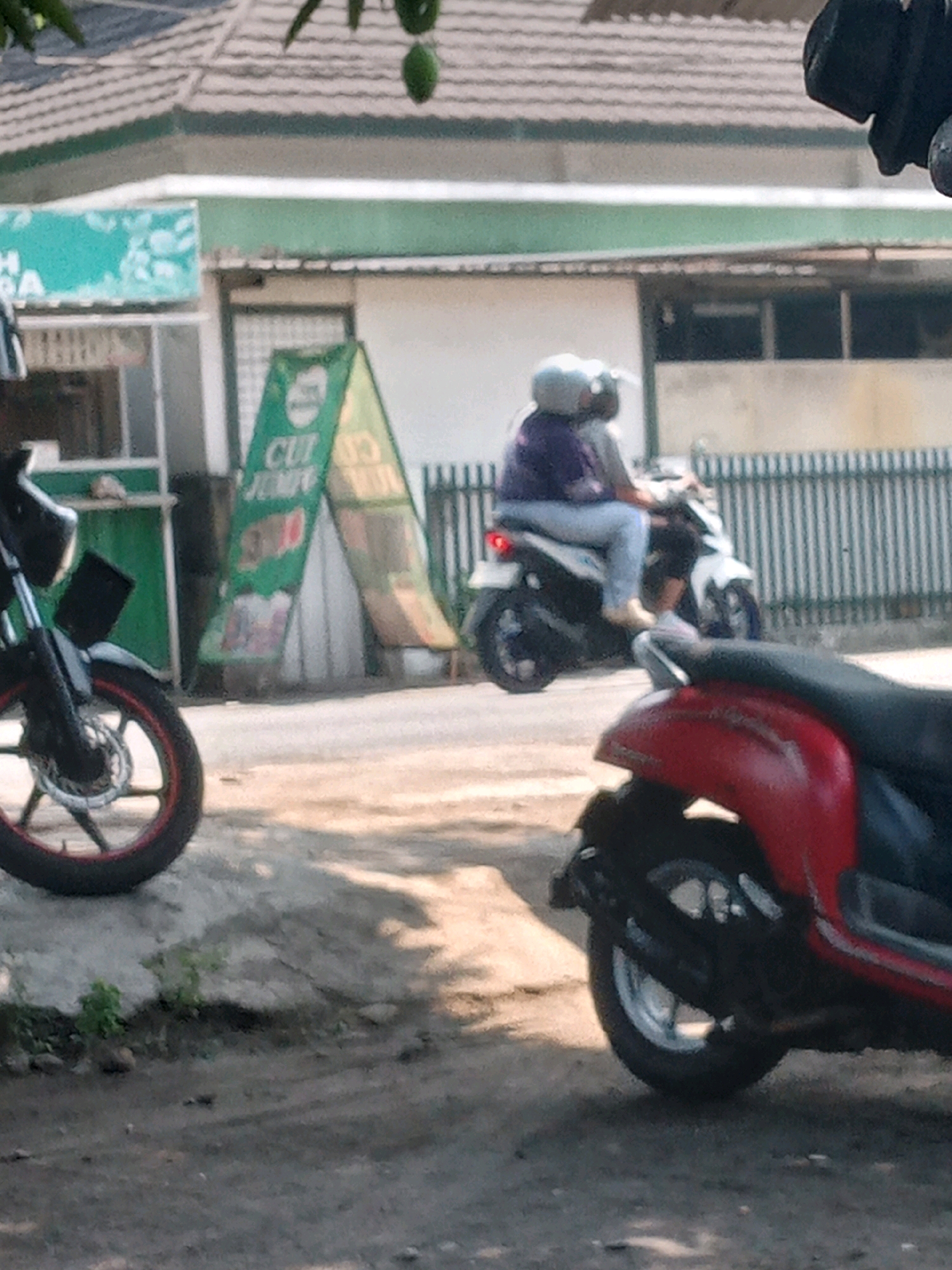 In Banguntapan, Indonesia, on a sunny day, two individuals ride a white and black motorcycle down a street. The rider in the front wears a white helmet and a light grey shirt, while the passenger behind wears a white helmet and a purple shirt. The motorcycle has a white body with black accents and a black seat. The street is paved with asphalt, and a wooden fence with vertical slats lines the right side of the road. A green and white advertising stand is visible on the left, with the text "CUT JUICE" printed on it. In the foreground, the front wheel of a black motorcycle with a red rim is visible, partially obscuring a red scooter on the right side of the frame. The background features a building with a tiled roof and several windows. Trees with green leaves hang over the scene, with two green fruits visible at the top.