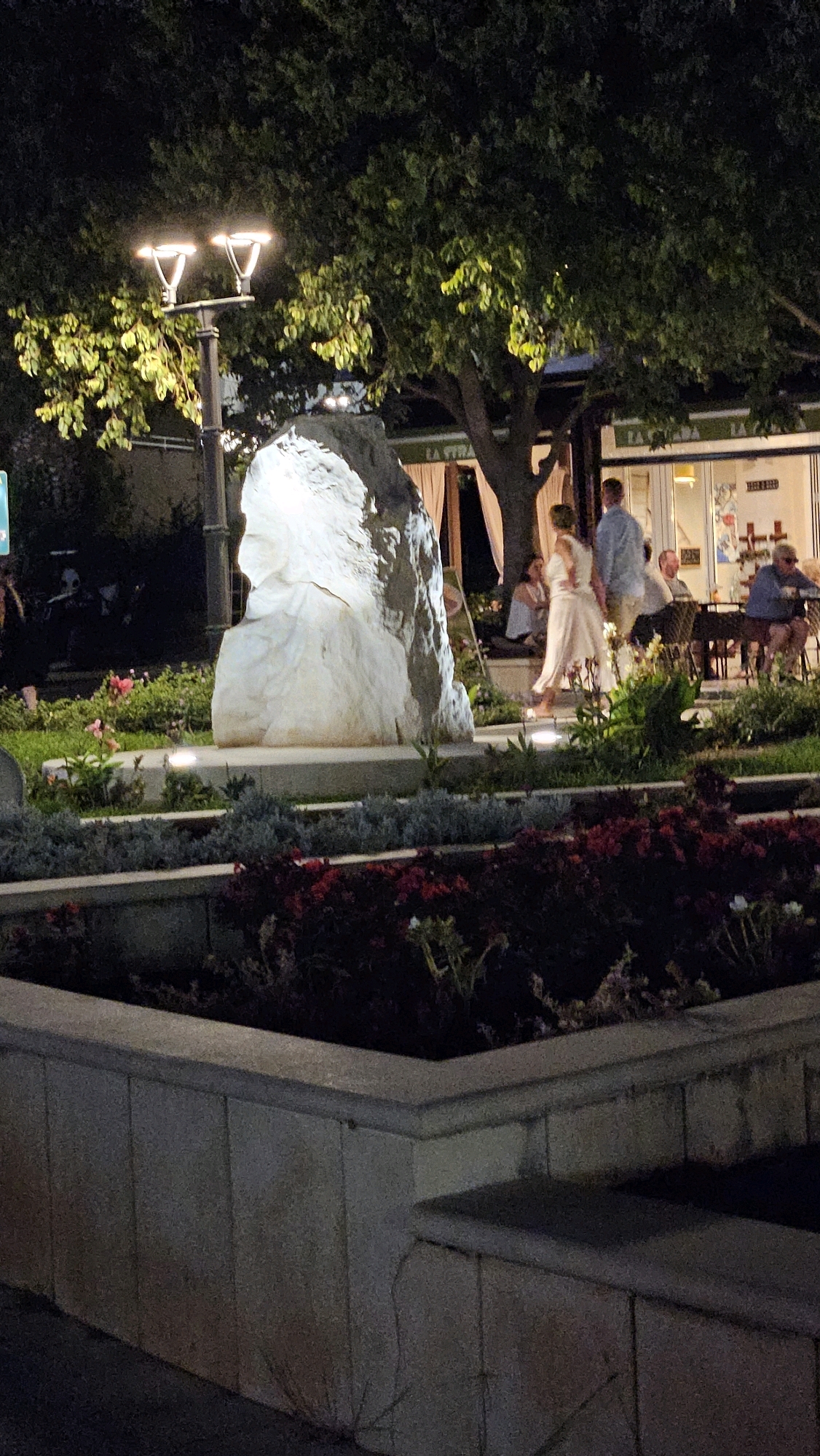 The image depicts an outdoor scene at night, likely in Općina Tučepi, Croatia. In the foreground, a raised planter box with dark red and purple flowers is visible. Behind the planter box is a large, light-colored rock sculpture, possibly abstract or depicting a figure. A lamppost with two lights stands to the left of the sculpture. In the middle ground, several people are seated at tables, seemingly at an outdoor restaurant or cafe. A couple is walking in front of the sculpture; the woman is wearing a light-colored dress, and the man is wearing a blue shirt. A large tree with green foliage extends over the scene. The background shows the facade of a building with awnings and windows. The overall atmosphere suggests a public area with dining and recreational spaces illuminated at night.