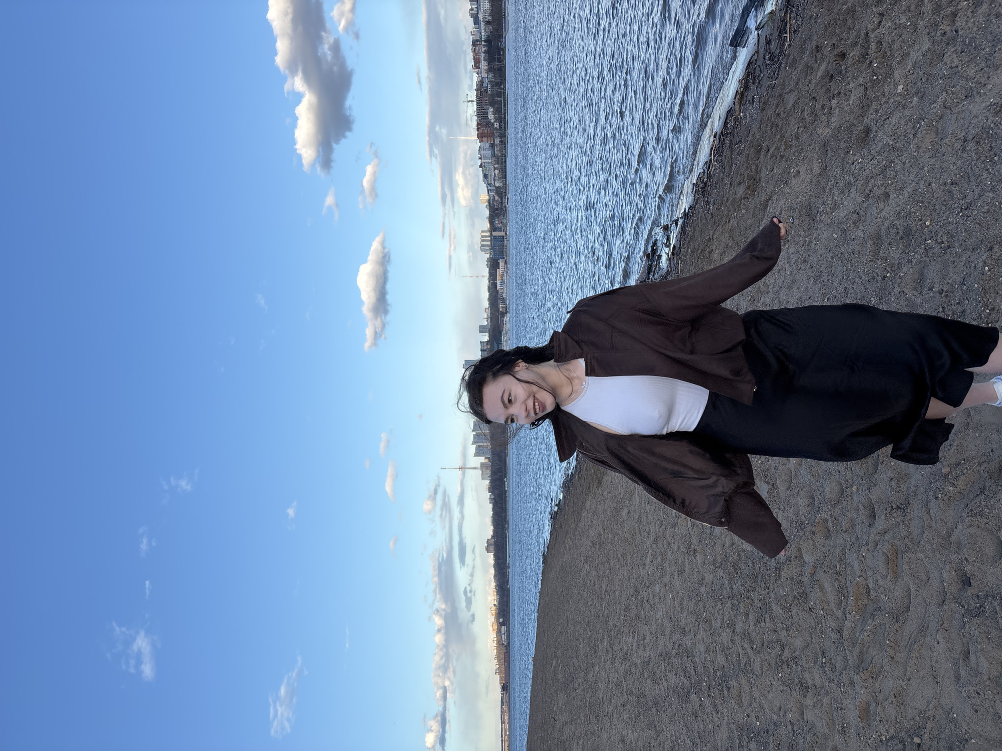 The image, correctly oriented, depicts a young woman standing on a sandy or gravelly shore next to a wide body of water, likely the Kama River in Perm, Russia. She is positioned towards the right side of the frame, looking towards the left and smiling broadly.
The woman is dressed casually in a brown, long-sleeved jacket worn over a white tank top, paired with a long black skirt and white sneakers. Her dark hair appears to be pulled back loosely.
In the background, across the expansive, rippled water, a city skyline is visible with various buildings and structures, suggesting an urban environment. The sky above is a clear blue with scattered white and light gray cumulus clouds, indicating fair weather. The light suggests it is late afternoon or early evening, with a soft, natural illumination on the scene. The sandy bank beneath her feet shows some footprints. No visible text is present in the image.