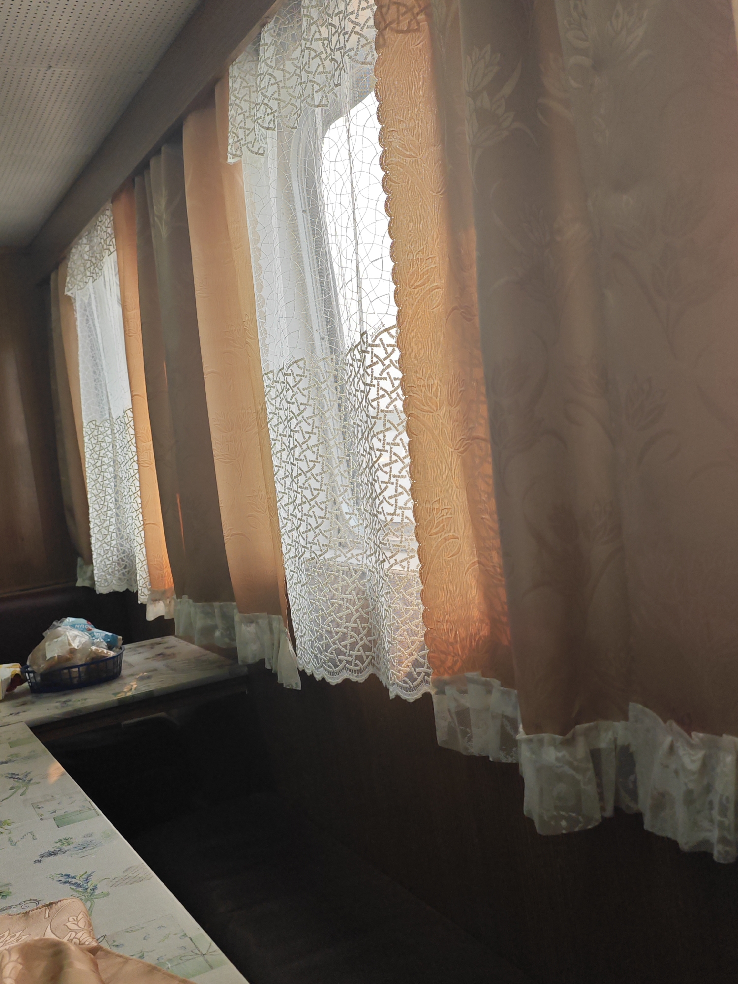 The image shows the interior of a room with curtains covering a window. The curtains are made of two layers: an inner layer of white lace with a geometric pattern and an outer layer of peach-colored fabric with a floral pattern. The lace also has a scalloped edge. The outer curtains are drawn, allowing light to filter through. There is a table in the foreground with a basket containing some food items, possibly bread. The table is covered with a patterned tablecloth. The room appears to be dimly lit, with sunlight casting a warm glow on the curtains. The overall impression is one of a cozy and private space. Without specific landmarks or identifying features, it is not possible to determine the exact location within Krasnoyarsk, Russia.