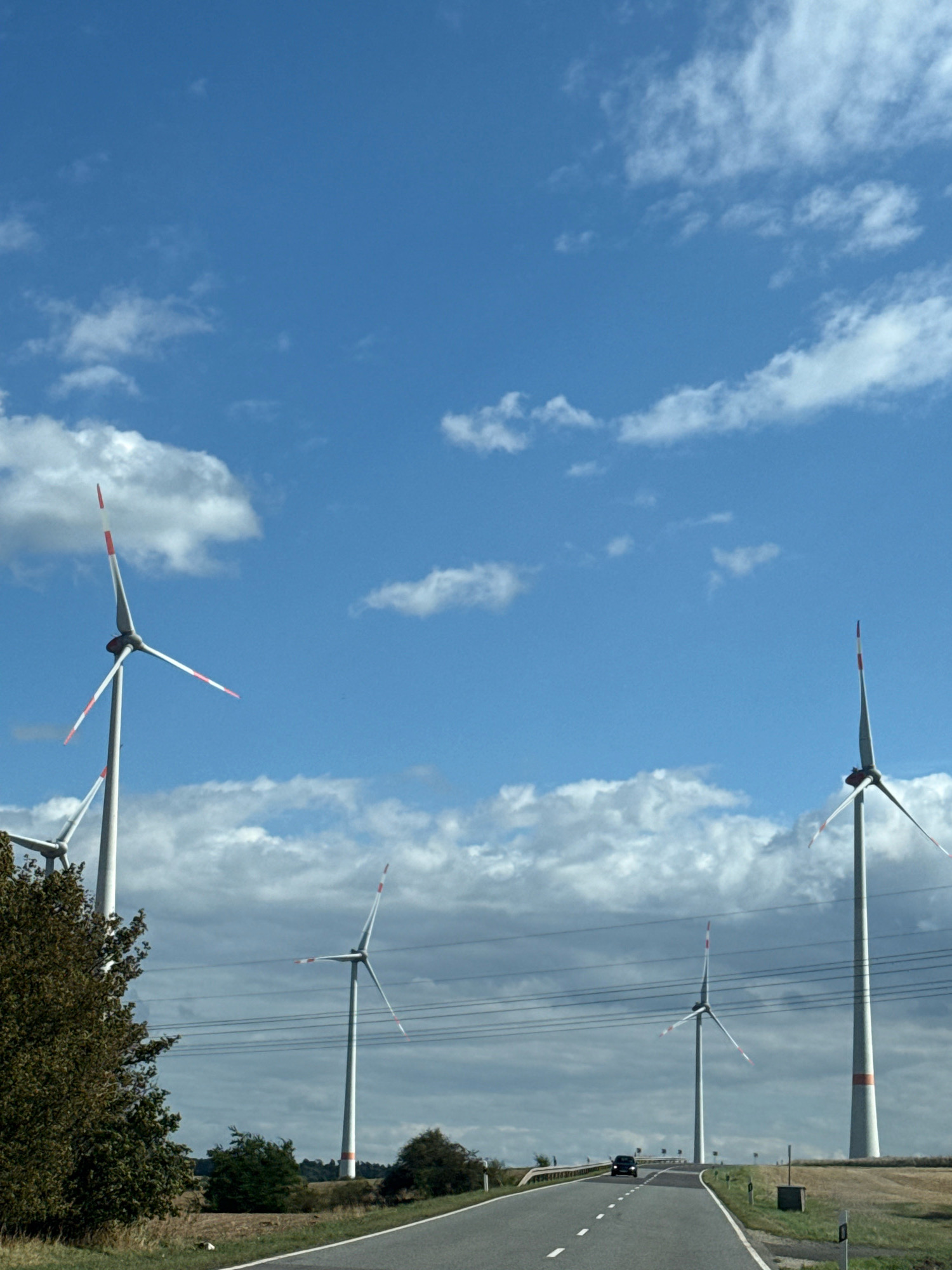 The image depicts a roadside view of several wind turbines under a partly cloudy blue sky. The scene appears to be located in Kleinfurra, Germany, given the presence of wind turbines, which are characteristic of the region's energy production landscape. In the foreground, a paved road curves to the left, with white dashed lines marking the lanes. A dark-colored car is visible driving on the road. To the left of the road, a large tree with dark green foliage stands prominently. Several tall, white wind turbines with red and white tips on their blades are spaced across the landscape, with some appearing larger and closer than others. Power lines are strung between poles in the middle ground. The terrain is gently sloped, with fields and some smaller trees visible in the distance. The overall impression is one of a rural, modern energy landscape.