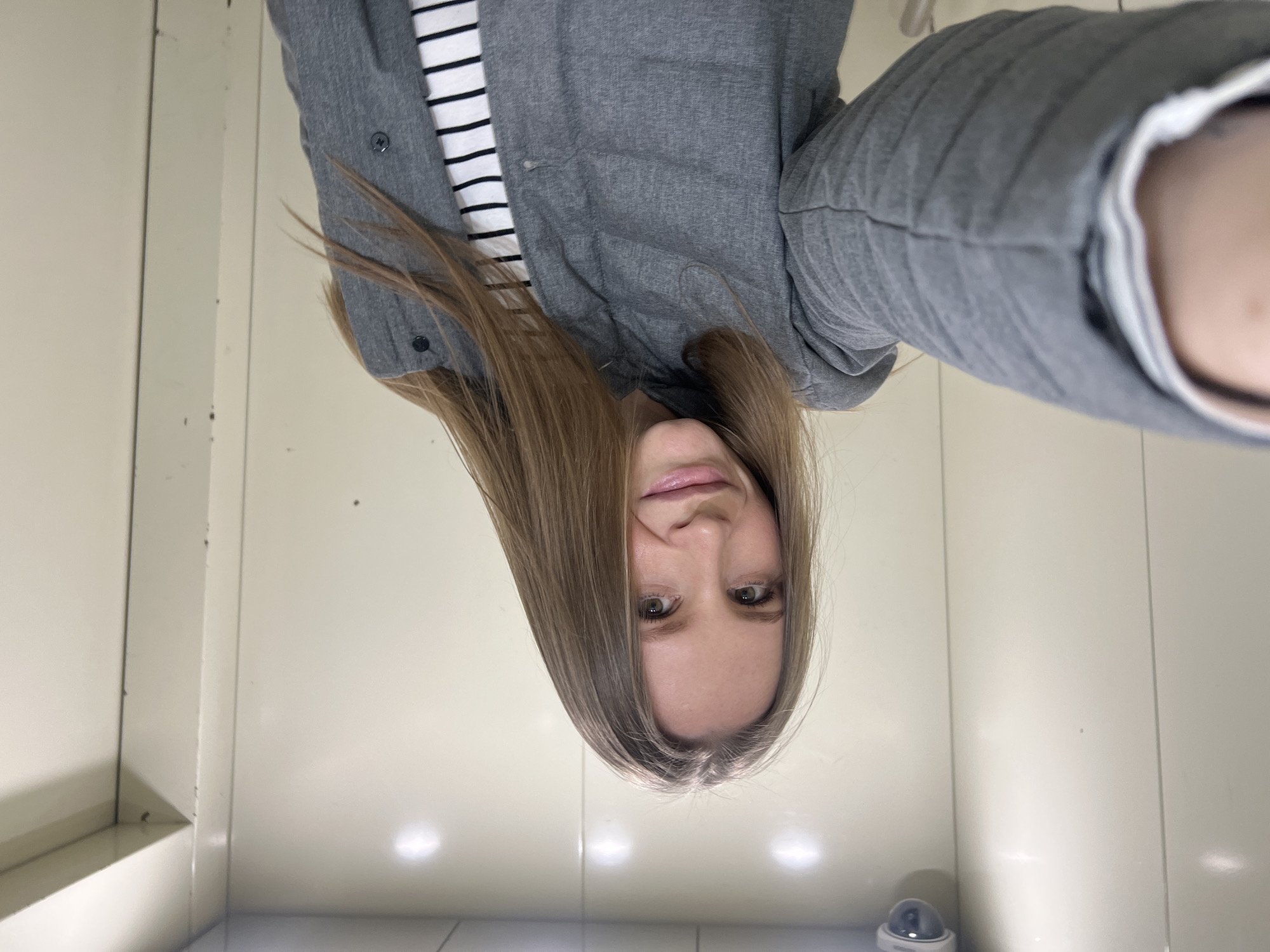 A woman with long, light brown hair is taking a selfie. She is wearing a gray jacket over a black and white striped shirt. She is in a small, enclosed space with white walls. A security camera is visible in the lower part of the image. The location is Khimki, Russia.