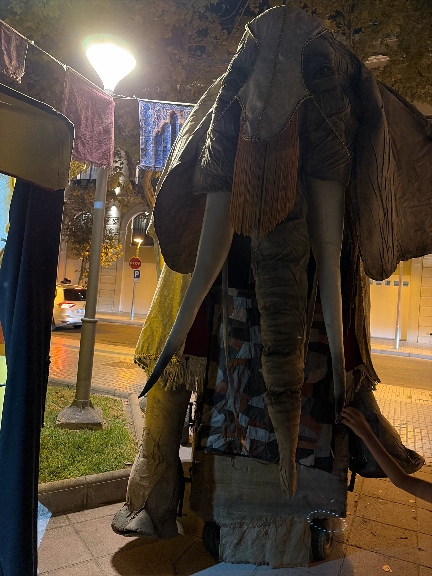 In the city of Salou, Spain, a large, elaborate puppet resembling an elephant stands on a wheeled platform at street level. The puppet is covered in fabric with a gray, wrinkled texture for the head and trunk, and it has long, off-white tusks. Draped over the puppet is a dark, textured fabric that extends outwards, resembling large ears or wings. A fringe of rust-colored material hangs from beneath the head. The elephant's legs are covered in a patchwork fabric of geometric shapes in brown, gray, and black. The puppet is situated in front of a building with yellow walls and arched doorways, illuminated by streetlights and an overhead lamp. In the background, a white car is parked on the street, and a red stop sign is visible on a pole. A small section of green grass is to the left of the puppet. A child's arm is reaching towards the right side of the puppet, interacting with its fabric covering. The overall scene appears to be an outdoor display or performance at night.