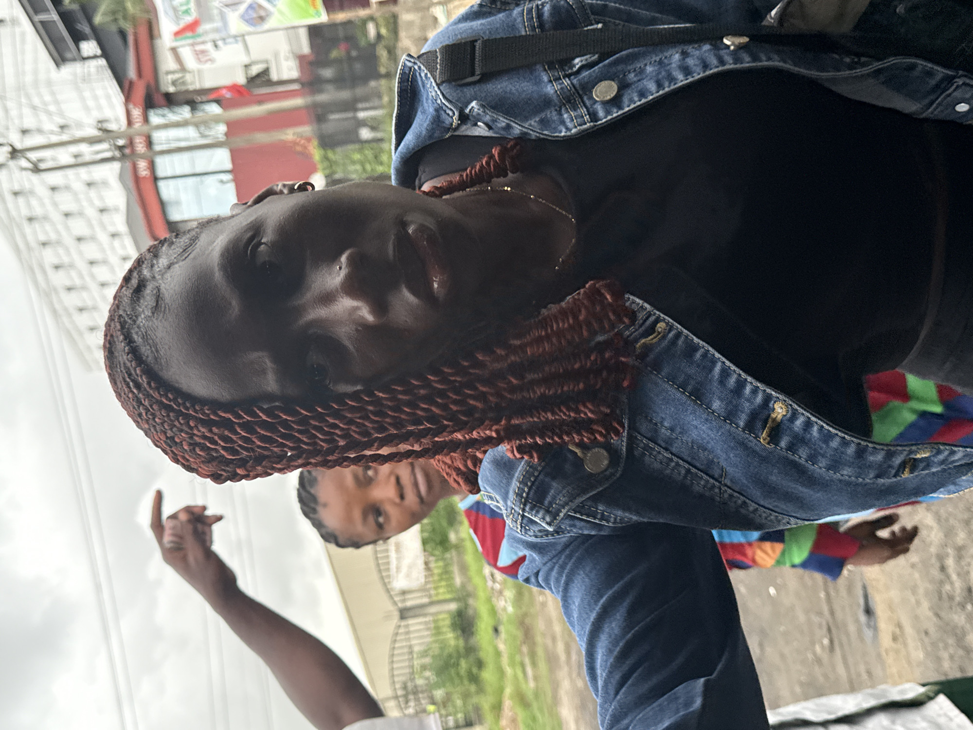 This outdoor medium shot features a primary subject, a dark-skinned woman with reddish-brown box braids or twists that cascade over her shoulders. She is wearing a blue denim jacket over a dark top. A thin silver chain is visible around her neck, and she has a small nose piercing and a hoop earring in her left ear. She looks directly at the camera with a calm expression and a slight smile.
In the mid-ground, to her right, another dark-skinned woman is partially visible, wearing a colorful top with blocks of red, blue, and green. Her gaze is directed slightly downwards or away from the camera. On the left side of the frame, a dark-skinned arm is raised, with the index finger pointing upwards; the rest of this person is out of view.
The background suggests an urban setting under an overcast sky. A tall, light-colored building with numerous windows is visible on the left. To the right, behind the second woman, is a building featuring red elements, possibly a commercial establishment. Partially visible, inverted text, including letters like "SW" and "AL," can be seen on a sign on this red building. Fencing and some green foliage are also present in the background. The scene depicts a daytime environment in a city.