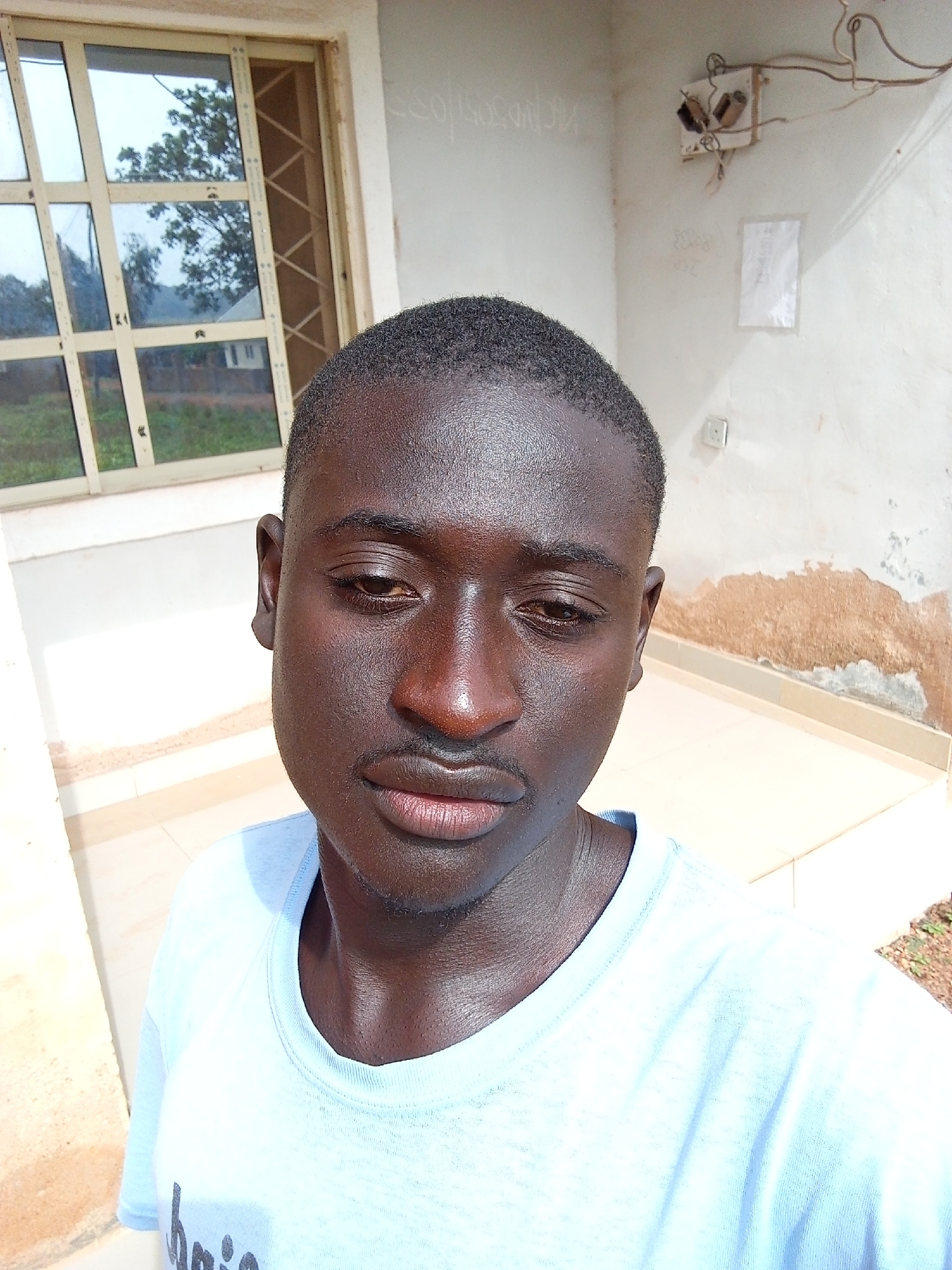 The image is a close-up selfie of a dark-skinned male with short hair, likely in his late teens or early twenties. He has a light blue t-shirt on. The background consists of a building exterior, featuring a window with multiple panes reflecting an outdoor scene of greenery. The wall appears to be partially peeling and has an electrical outlet with wiring exposed. There is also a white paper attached on the wall next to the outlet. The scene appears to be Bukuru, Nigeria, based on the request for location context.