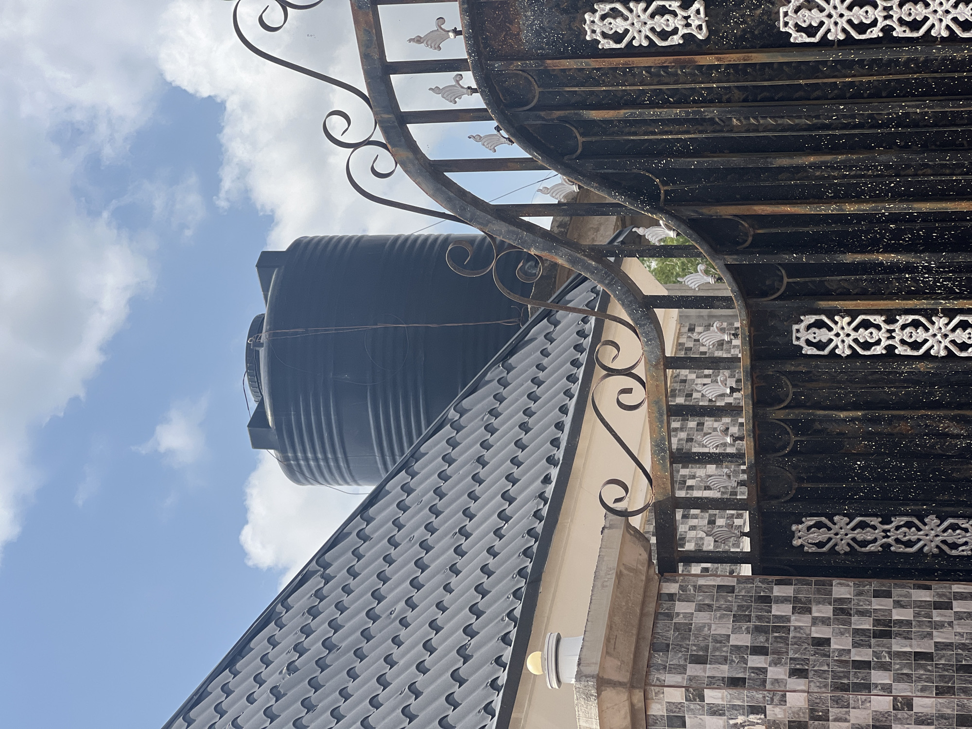 The image displays a low-angle view of a black water tank situated on the roof of a building, which has a dark, patterned roof. The building is adorned with patterned tiles and appears to be partly covered by an ornate black metal gate. The gate features decorative scrollwork and white filigree details. In the background, a bright blue sky with scattered white clouds suggests it is daytime with fair weather. The overall scene is of an exterior view of a building, possibly in a residential area in Howayi, Nigeria. No people or specific activities are depicted, and there is no visible text in the image. The lighting indicates it is daytime, likely sunny, given the brightness of the sky and the clear visibility of the objects.