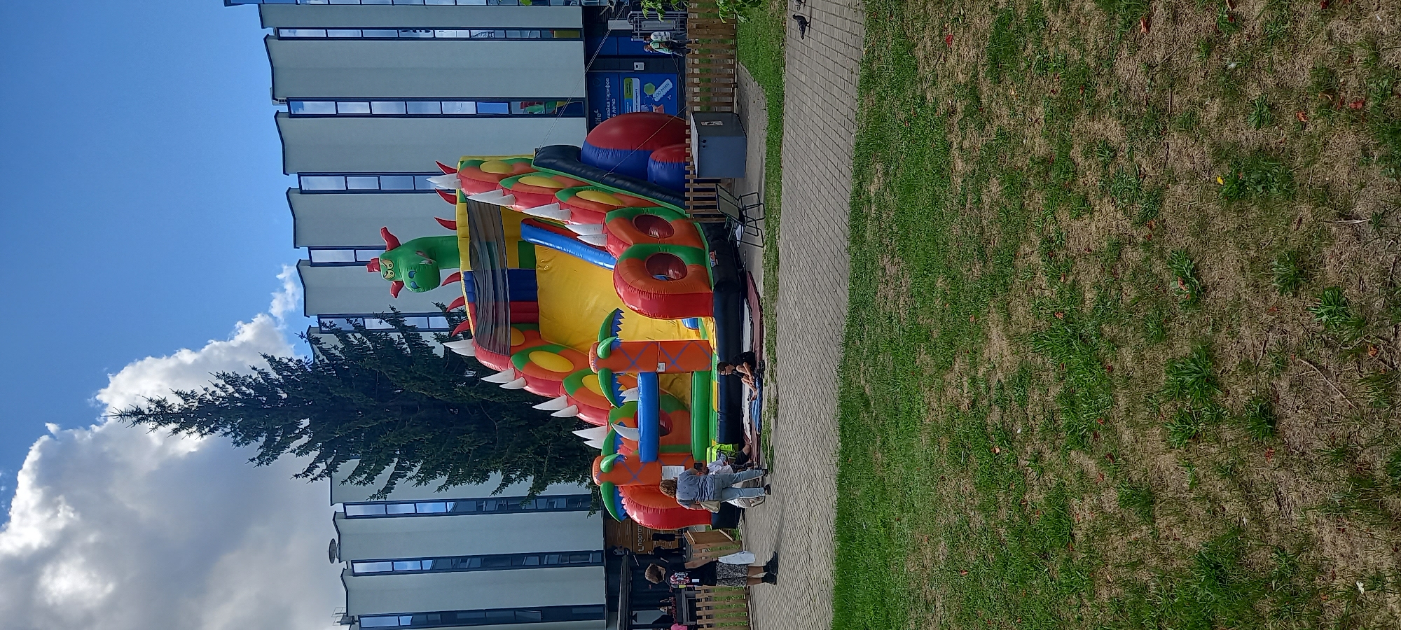 The image shows a vibrant, inflatable dragon-themed playground structure situated on a paved area adjacent to a grassy lawn. A tall evergreen tree stands behind the inflatable, with a modern building featuring a distinctive facade of vertical white panels and blue-tinted windows as the backdrop. The sky is bright blue with scattered white clouds.
In the foreground, on the paved area, a person in a black t-shirt and shorts stands with their back to the camera, holding a white bag. To their right, a woman in a blue shirt and jeans faces the inflatable, holding a tote bag. A child is seated on the ground near the inflatable. Another person is also sitting on the ground to the right of the woman, wearing a black shirt and shorts.
The inflatable playground is a prominent feature, designed like a dragon with a green head at the top, red and orange spikes, and a yellow slide. It is colorful, with sections of red, orange, green, and blue. The structure appears to be set up for children's entertainment.
In the background, the modern building has a clean, linear design. A wooden fence runs along the edge of the paved area. A grey waste bin is visible near the fence. On the building, there are signs, one of which includes Cyrillic text and what appears to be a logo. A few pigeons are visible on the paved ground.
The overall scene suggests an outdoor recreational area in a city. Based on the Cyrillic text on the signage visible in the image, it is highly probable that this location is in Brest, Belarus, a city where Cyrillic is the primary script. The presence of the inflatable playground and the general urban setting indicates a public space designed for leisure activities, likely during daytime hours given the bright lighting and clear sky.