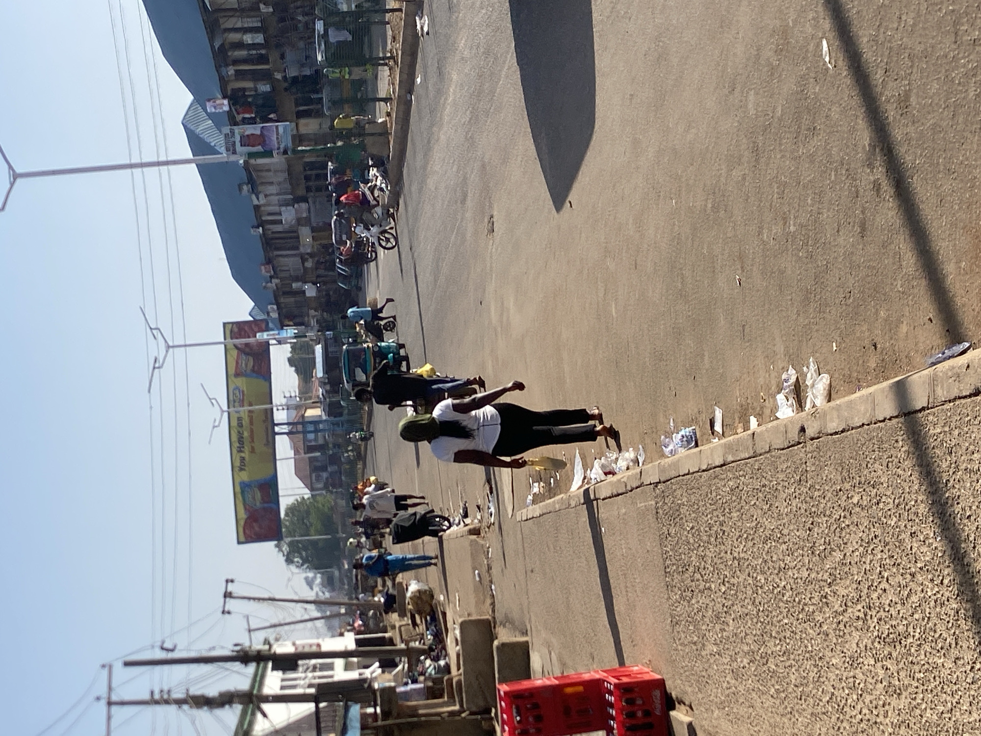 The image depicts a street scene in Bukuru, Nigeria. A woman in a white t-shirt, black pants, and a green head covering is walking alongside the road. Litter is scattered along the curb. In the background, there are several motorcycles and people, including some selling goods. A large billboard is visible over some shops. There is a row of shops running along the side of the road, with a metal roof over them. Overhead power lines can also be seen. A red plastic crate is on the near left. The street is relatively empty and there are shadows cast by the sun.