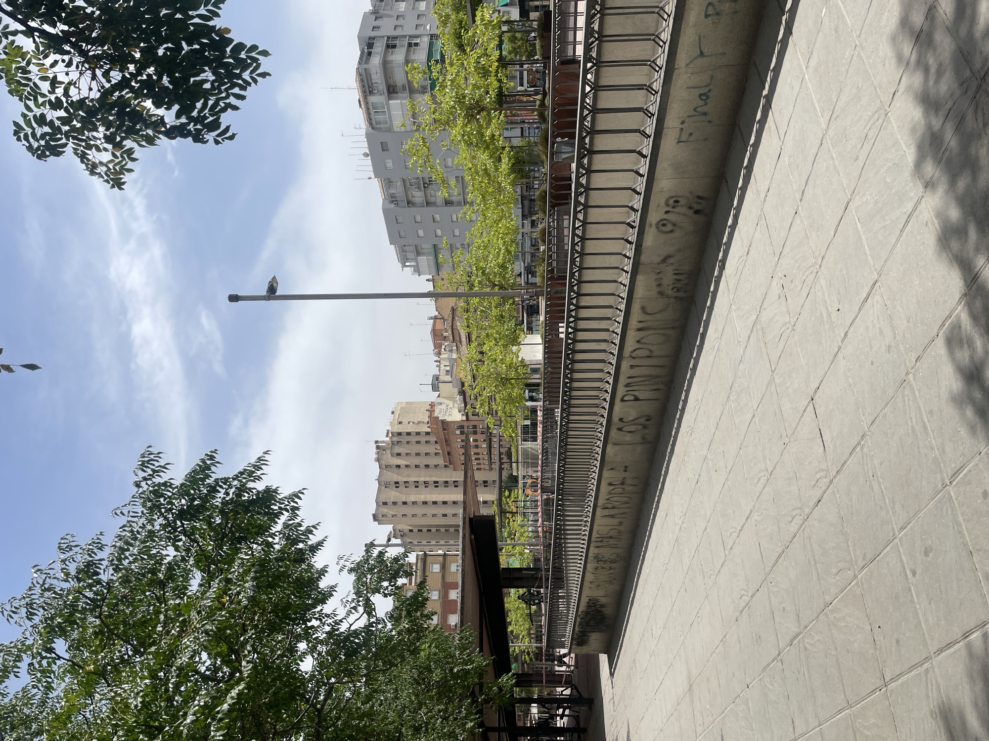 This image depicts an outdoor urban scene in Granada, Spain, specifically the area around the Plaza de Bib-Rambla, looking towards Calle Elvira. In the foreground, a light-colored tiled pedestrian walkway stretches across the frame, casting long shadows from an unseen source of light. A low concrete wall with graffiti runs along the middle ground, topped with a black metal railing. The graffiti appears to be in Spanish, with phrases like "LOS PIN PON" visible.
Beyond the wall, a covered open-air structure with dark supports and a brown roof is visible on the left. This area seems to be a public space, possibly a market or seating area, with some people walking in the background. Further into the midground and background, several trees with vibrant green foliage are planted in rectangular planters, interspersed with a street lamp.
The background is dominated by a series of multi-story buildings of varying architectural styles. On the left, a tall, beige building with numerous windows stands out. To its right, several other buildings with different colored facades and rooflines are visible. In the center and right of the frame, more modern, grey apartment buildings with balconies are prominent. The sky above is a light blue with scattered white clouds. The overall impression is that of a sunny day in a lively urban square.