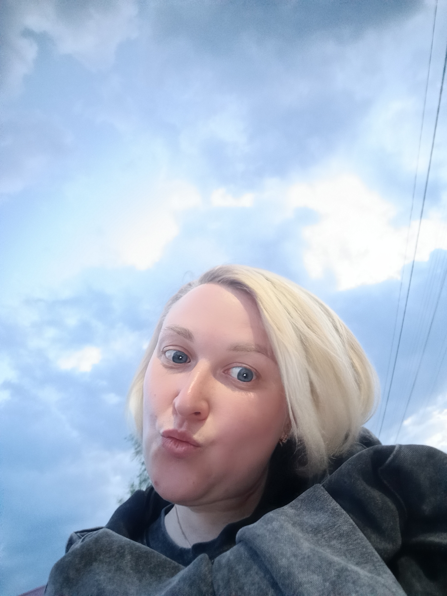 A selfie of a woman with blonde hair and blue eyes. She is wearing a dark gray sweatshirt. She is making a kissing face. The background shows a cloudy sky with power lines. The exact location is impossible to determine from the image alone. The request for the location to be Chaikovsky, Russia cannot be confirmed based on visual evidence.