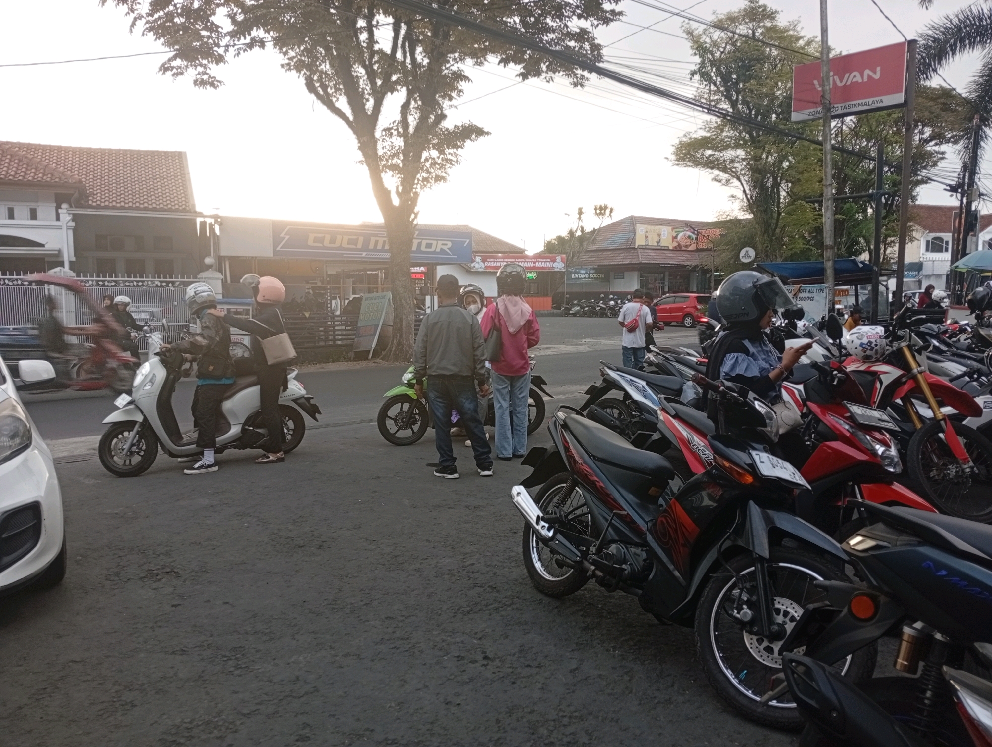 The image depicts an outdoor scene in Tasikmalaya, Indonesia, likely a street or parking area. The lighting suggests it is either early morning or late afternoon. Several motorcycles are parked or in motion, with some people on them. In the foreground, a white car is partially visible on the left. Several individuals are seen on or near motorcycles. One white scooter is occupied by two people wearing helmets. Further back, a green motorcycle is parked, and a few people are standing near it. In the mid-ground, a red and black motorcycle is prominent, and behind it, a row of other motorcycles, including red and white ones, are parked. A red sign with the word "VIVAN" is visible in the background on a pole, along with text below it that appears to indicate a location within Tasikmalaya. Buildings with storefronts are visible on the left and in the mid-ground, suggesting a commercial area. Trees are present, with some foliage casting shadows. Power lines are strung across the sky. The ground is asphalt with some visible wear and tear.