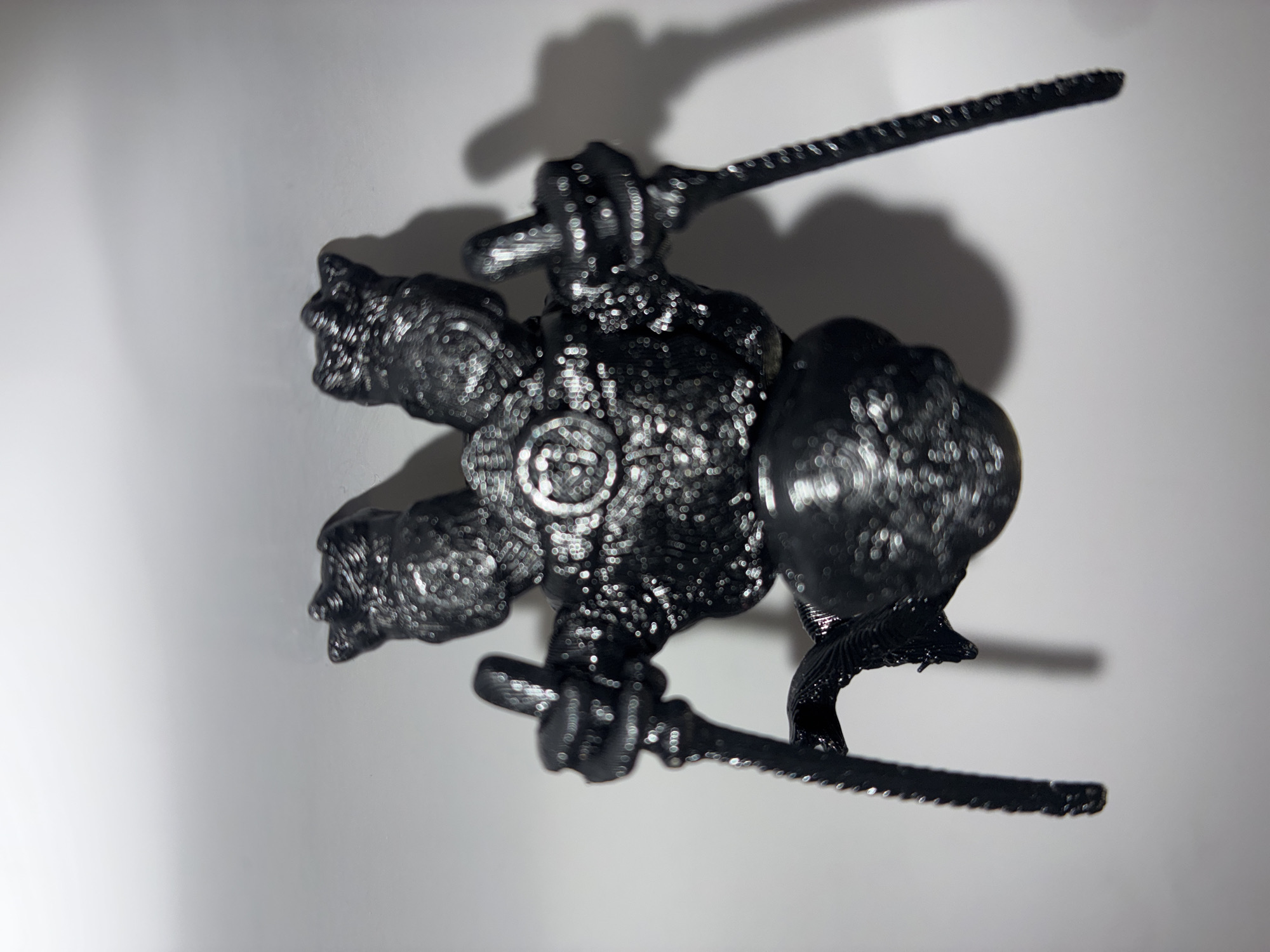 This is a close-up, top-down shot of a small, black, 3D-printed figurine. The figurine appears to be a stylized character, possibly a warrior or mythical creature, with multiple limbs and what looks like weapons, such as swords or scythes, extending from its body. The details of the figurine are intricate, showing layered textures indicative of the 3D printing process.
The figurine is placed on a white, flat surface, which could be a table or a backdrop. A soft shadow of the figurine is cast to the upper right, suggesting the light source is positioned above and to the left of the object. The lighting creates a slightly diffused effect, with some highlights visible on the glossy surface of the figurine.
The focus of the image is on the figurine itself, with the background being largely out of focus. There is no discernible text or writing visible on the figurine or its surroundings. The scene is simple and lacks specific environmental cues that would indicate a particular time of day, weather, or outdoor location. The context provided suggests the photograph was taken in Toruń, Poland, but this information is not visually evident within the image itself. The overall impression is of a product photograph or a personal item being showcased.