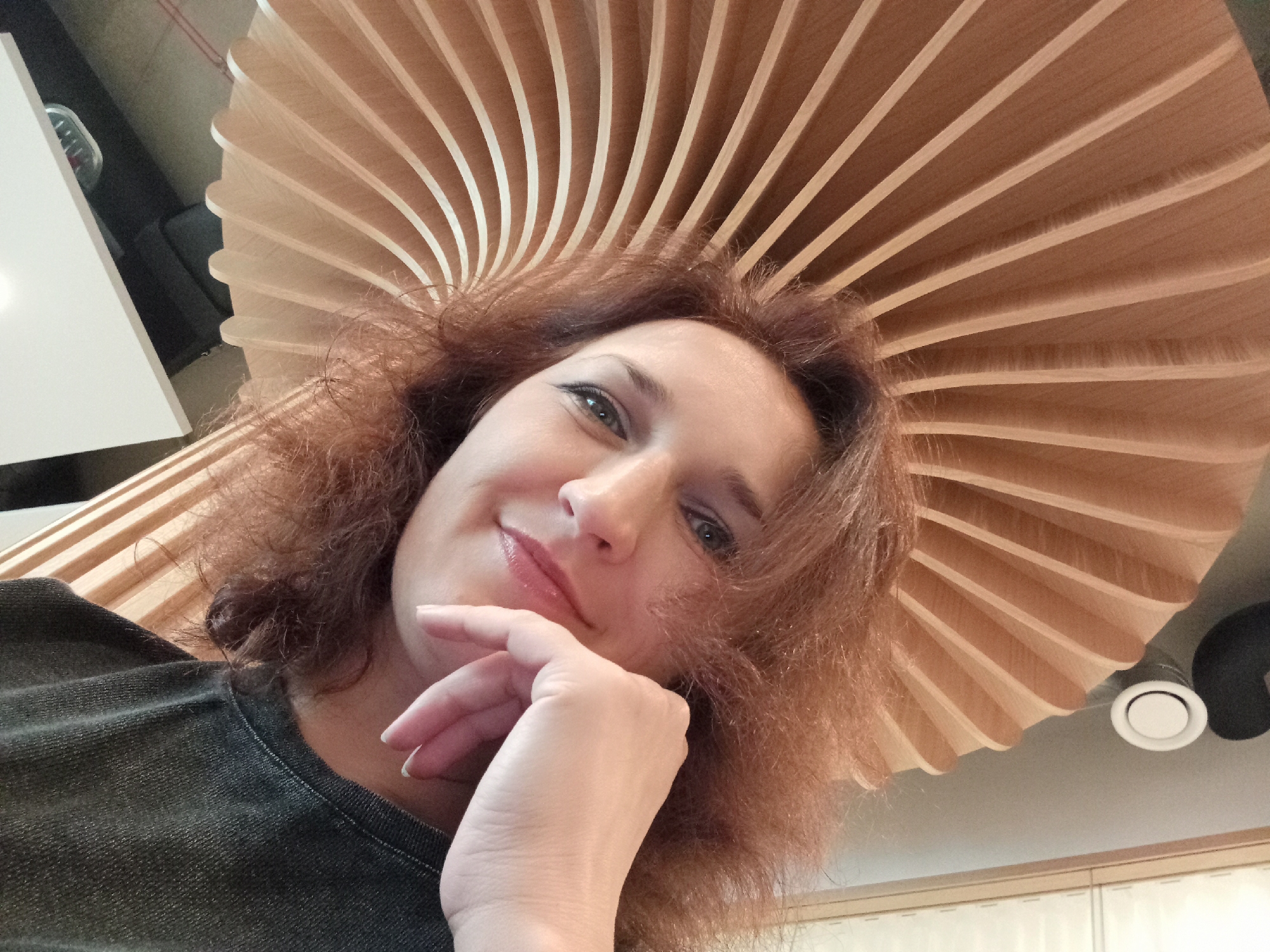 A medium shot is captured from a low angle of a woman looking up at the camera. She has reddish-brown, curly hair, and is wearing a dark grey t-shirt. Her right hand is raised, and her fingers are gently resting on her chin. She is smiling, with her lips slightly parted, and her eyes are looking directly into the camera lens. Her makeup includes eyeliner and lipstick. Behind her, a wooden ceiling installation resembling a fan or umbrella is visible. The installation has many thin, curved wooden slats radiating outwards. The environment appears to be indoors, possibly in a modern building. The lighting is bright, illuminating the woman's face and the wooden structure. There are no visible landmarks or signs to indicate a specific location in Riga, Latvia.