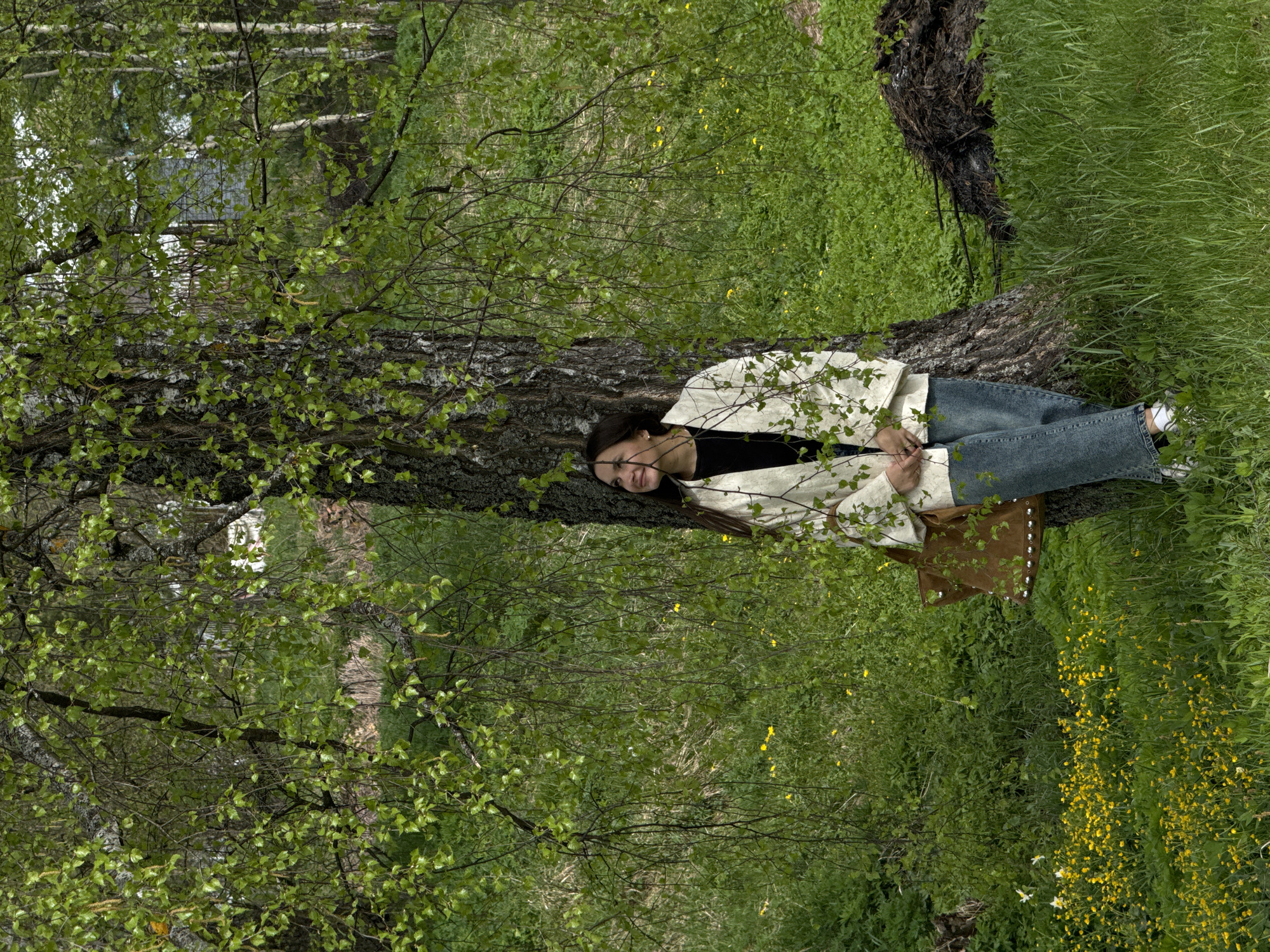 A woman is lying against a tree in a grassy area in Красный Маяк, Russia. She is wearing a light-colored jacket, a black shirt, and blue jeans. Her head rests against the trunk of the tree, and her body extends along the base of the trunk. A brown bag is next to her. The area is overgrown with green vegetation and small yellow flowers. The tree has new green leaves.