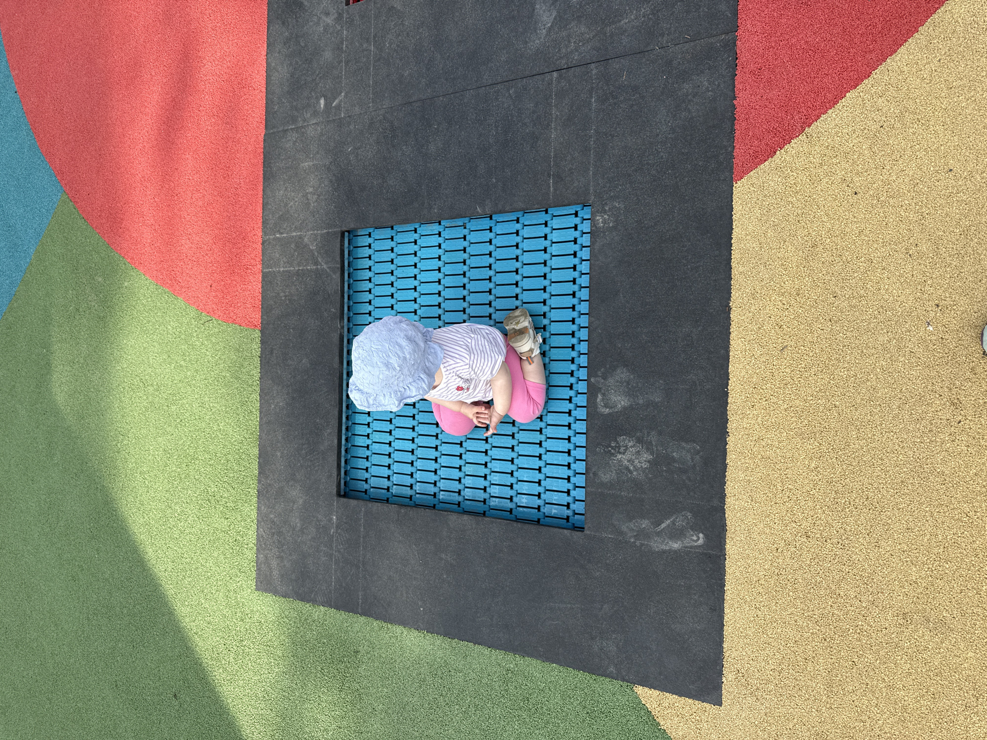 The image shows a child sitting on a blue springy square surface within a black square frame, part of a playground. The playground surface surrounding the square is made up of various colored sections: red, green, yellow, and light blue. The child wears a light blue hat, a striped shirt, pink leggings, and white shoes. The location might be a public playground in Moscow, Russia, but there are no specific indicators to confirm this.