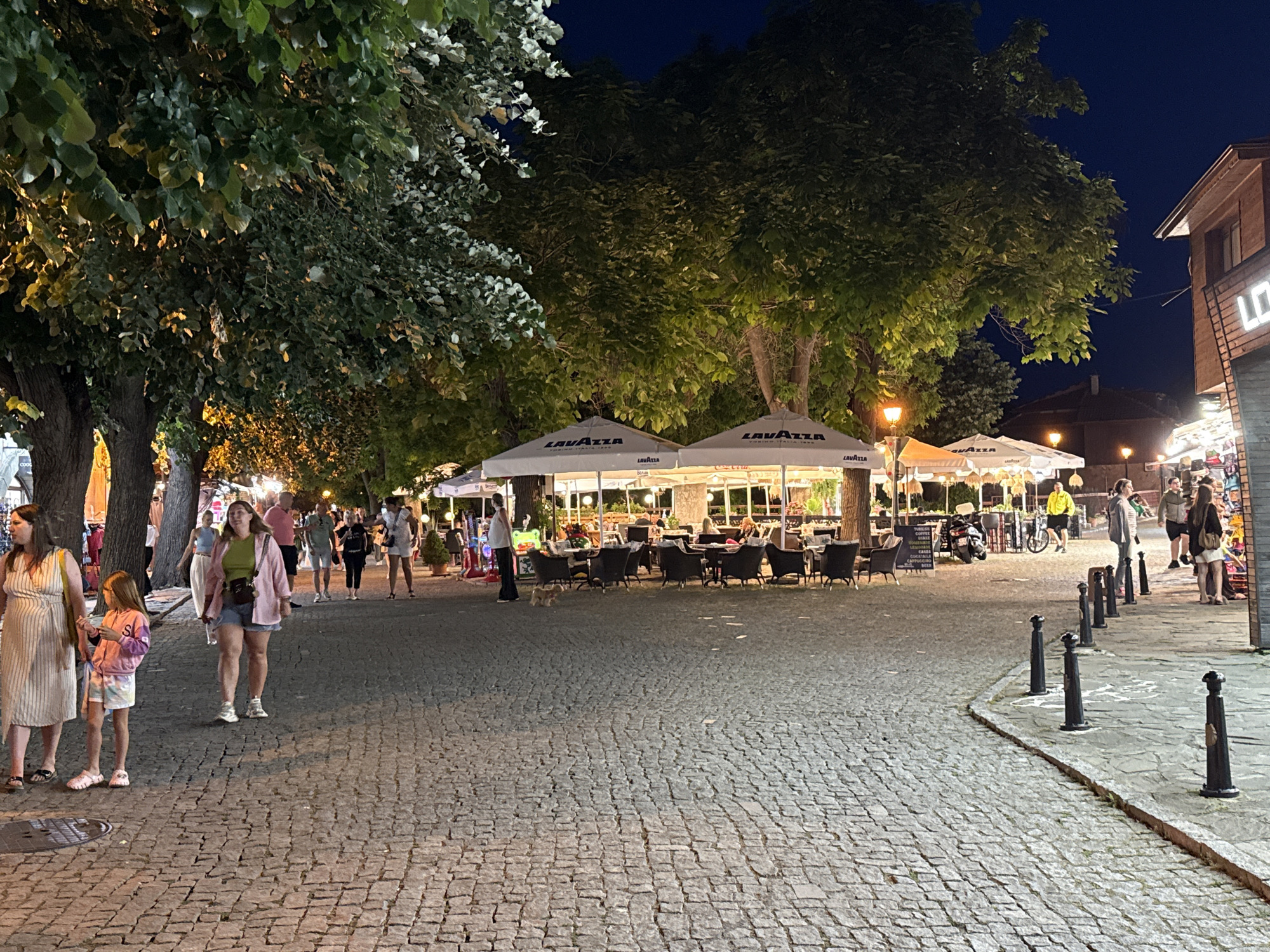 The image depicts a paved street scene at dusk in Nesebar, Bulgaria. The street is made of cobblestones and is lined with trees on the left, casting shadows and illuminated by streetlights. On the right side of the street, there are buildings with awnings and outdoor seating areas, suggesting restaurants or cafes. Several people are visible walking along the street or seated at tables. Some of the awnings have the "Lavazza" logo. There are also bollards lining the edge of the sidewalk. In the background, more buildings and trees are visible under a dark sky. The overall atmosphere is that of a lively evening in a European town.