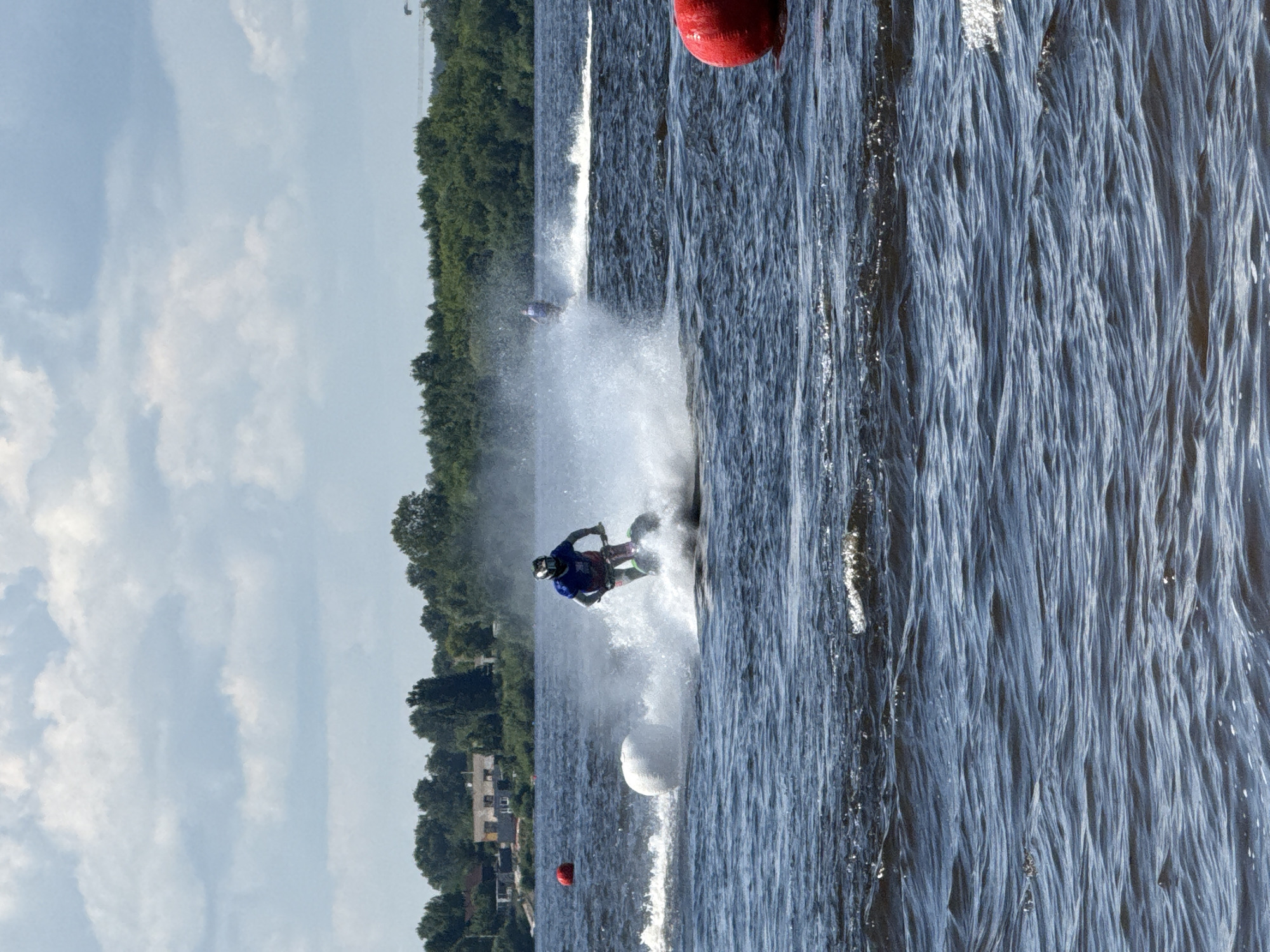 On a bright, cloudy day, a person on a jet ski is racing on a body of water, creating a large spray of water. The jet ski is dark green, and the rider is wearing a blue shirt and a black helmet. In the background, there is a line of green trees and some buildings. There are several buoys in the water; a large red one is in the foreground on the right, and two white buoys are to the left of the jet skier. There is also a red buoy in the background on the left. The jet ski is likely in the Kama River near the city of Perm, Russia, as the scenery is consistent with the riverbanks.