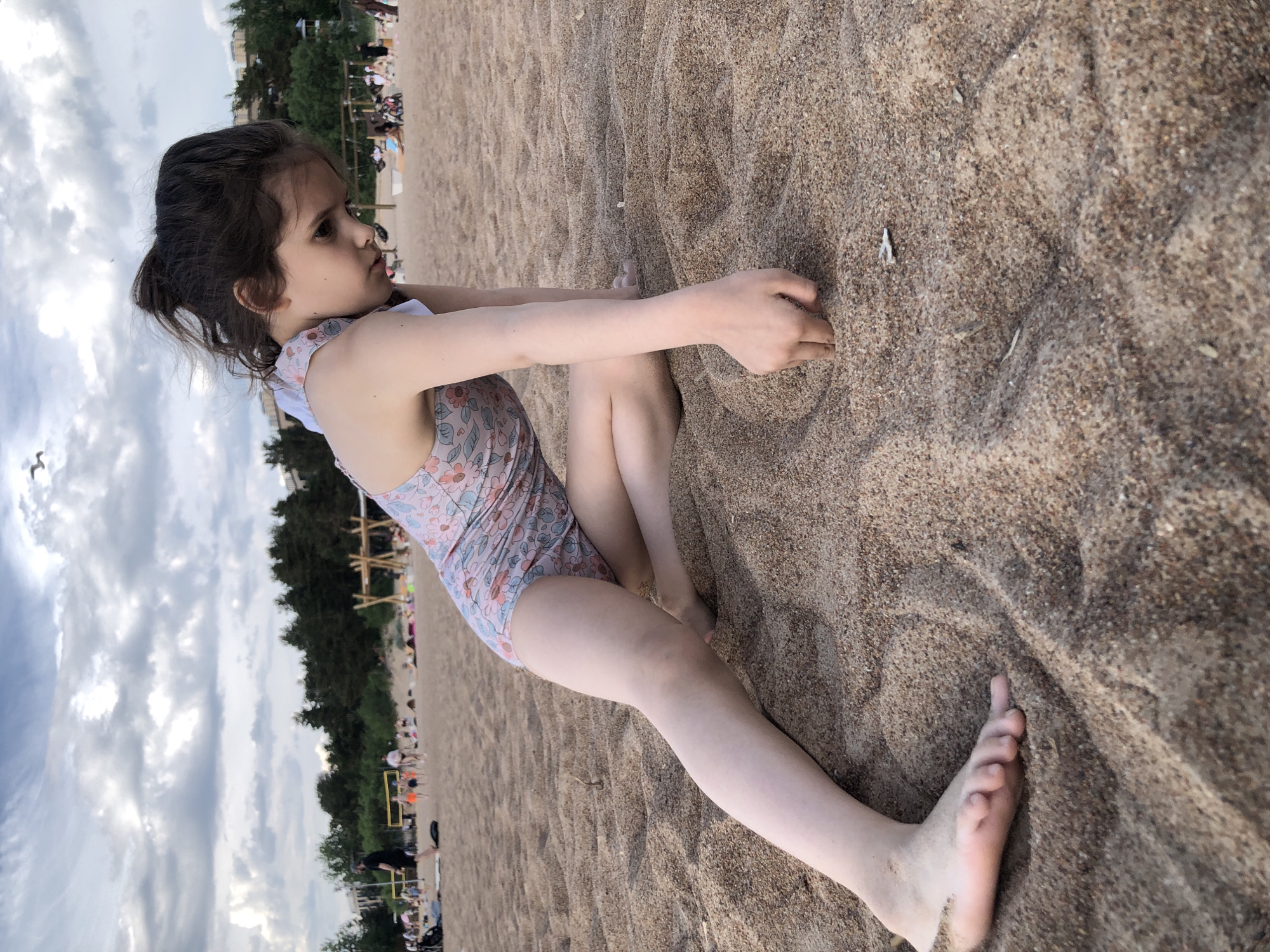 The scene depicts a sandy beach in Sestroretsk, Russia, under an overcast sky. A young female child, estimated to be between five and eight years old, with dark brown hair pulled back, occupies the foreground. She is wearing a light pink one-piece swimsuit adorned with a repeating pattern of small flowers and birds. The child is positioned on the light brown, fine-grained sand, leaning forward with her right arm extended, her fingers actively sifting or digging into the sand. Her left leg is bent, while her right leg extends forward along the sand, with her bare foot visible. Her gaze is directed downwards towards her hands and the sand.
In the midground, numerous indistinct figures of individuals are visible across the beach, engaged in various activities such as walking, playing, and resting. A yellow-framed volleyball net is present towards the left-center of the midground. To the right, a wooden playground structure, possibly a swing set, is discernible.
The background features a dense line of dark green coniferous trees, beyond which light-colored multi-story buildings are visible on the horizon. The sky is predominantly covered with a mixture of grey and white clouds. A single white bird is captured in flight in the upper right quadrant of the image. The sand surface exhibits subtle undulations and scattered small debris.