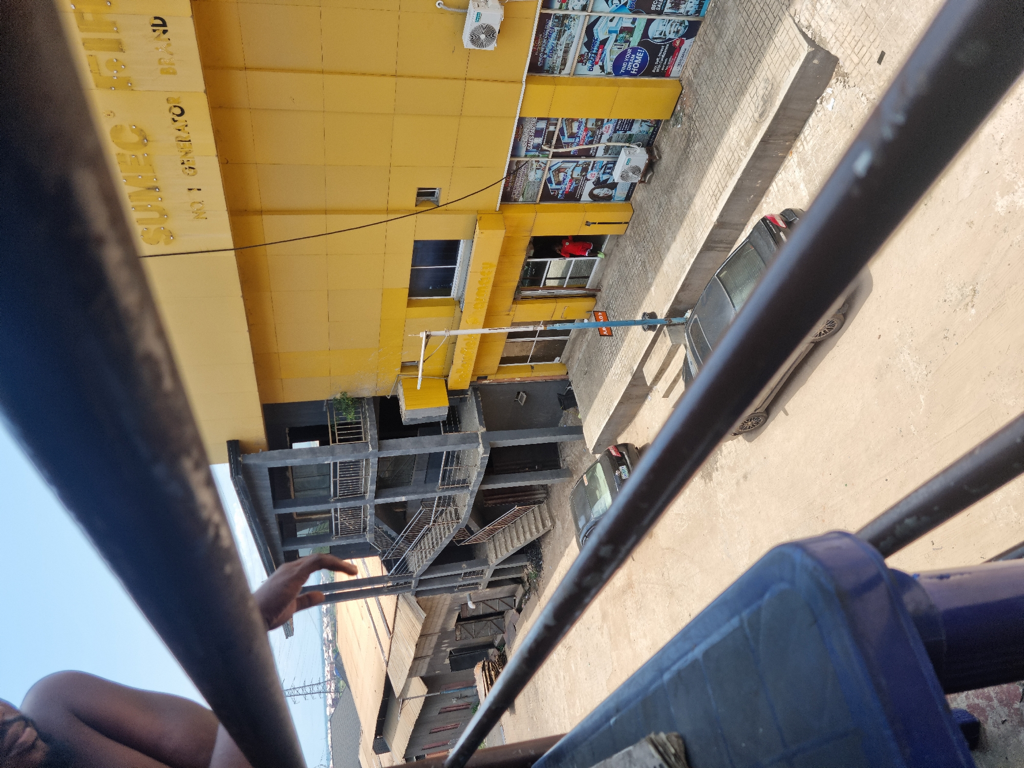 This image, taken in Onitsha, Nigeria, captures an urban street scene from a high, tilted vantage point, possibly from a vehicle or elevated structure, with dark metal railings crossing the foreground.
The central subject is a vibrant yellow multi-story building, likely commercial, with numerous dark-framed windows and several air conditioning units mounted on its exterior. Below it is another multi-story concrete structure with exposed stairs and railings, appearing industrial or under construction. On the street level, a dark-colored car is visible, parked or moving.
A person's arm and shoulder are visible in the immediate foreground on the left, next to a dark blue object. Another person in a red shirt can be seen partially inside a ground-floor doorway of the yellow building.
Notable details include the bright, sunny weather indicated by strong shadows. The perspective is downward-looking, creating a sense of height.
Visible text on the yellow building includes "SUMEC", "NO 1 GENERATOR", and "BRAND", suggesting a business related to generators. Posters and signs on the yellow building display phrases like "FIND YOUR DREAM HOME!", "SALES OFFICE", and "BEGANCOUVE". Other partially visible text includes "FIF" and "BEC".