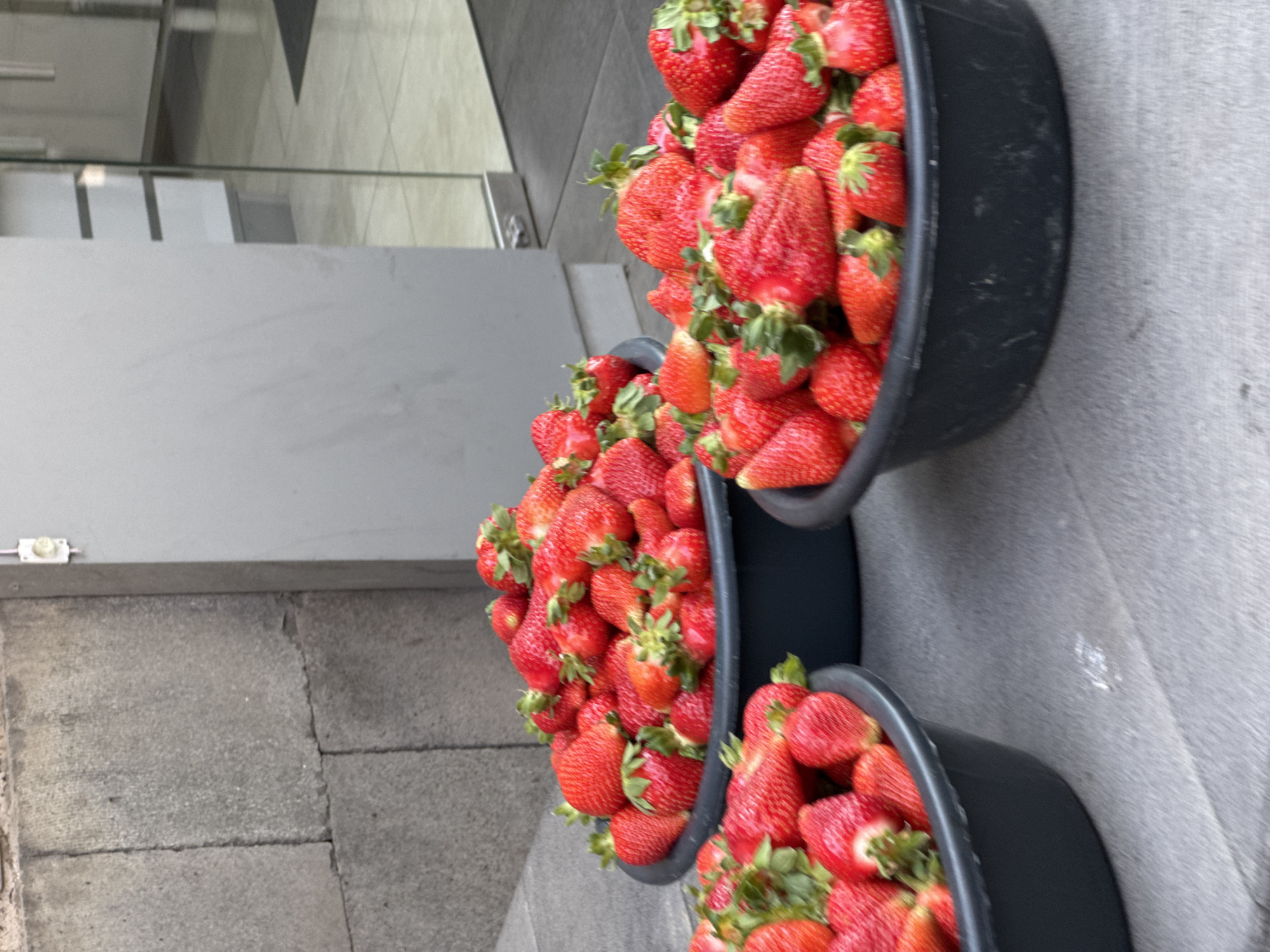 The image shows three black buckets filled with fresh red strawberries on a gray stone surface. The perspective is a close-up, angled downward towards the buckets. The buckets are stacked in a slightly staggered arrangement, with the largest bucket at the top and smaller ones below. The scene is lit by natural light. Details of a building with glass elements are visible in the background, suggesting an urban setting. It is not possible to confirm the location as Yerevan, Armenia, from the image alone.