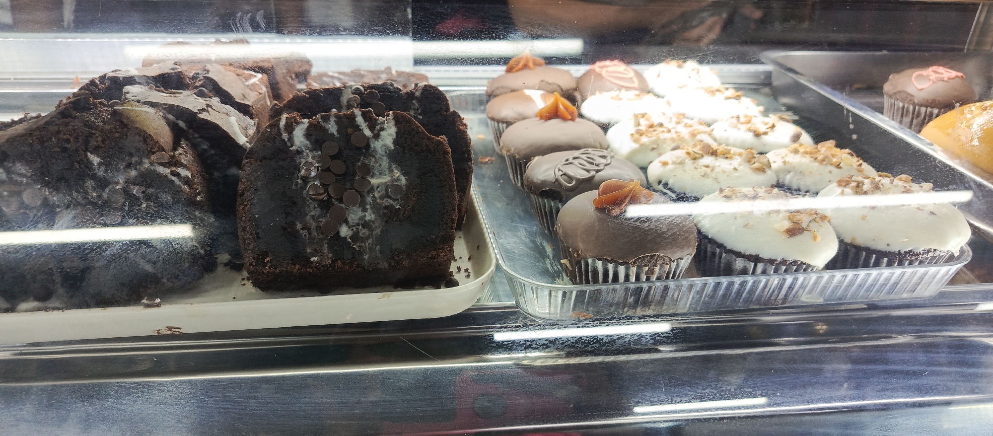 A display case contains desserts in Altagracia, Venezuela. On the left side, there's a tray of cut brownie-like desserts with chocolate chips and white frosting. Next to it, a tray filled with cupcakes, some with chocolate frosting and caramel topping, others with white frosting and chopped nuts. One cupcake has pink frosting. A glazed pastry is also visible. The case is behind glass.