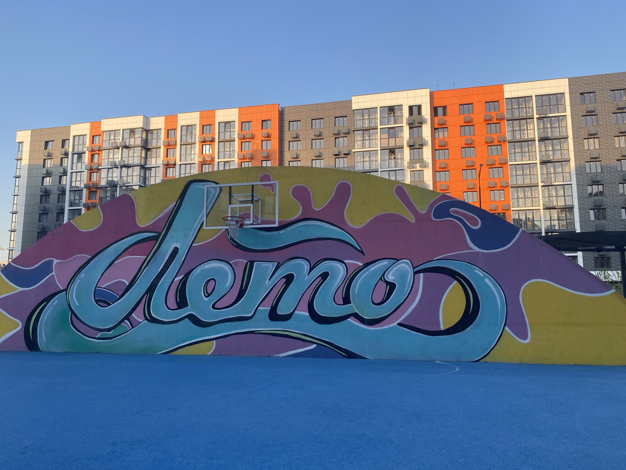 A basketball court with a vibrant mural is set against a backdrop of modern apartment buildings under a clear blue sky. The mural, painted on a curved wall, features the word "Лето" (Lieto, meaning "Summer" in Russian) in large, bubbly, teal-colored graffiti letters. The letters are outlined in black and white, with subtle shading that gives them a three-dimensional effect. Behind the lettering, the wall is painted with abstract splashes of pink, purple, yellow, and blue, creating a playful and energetic design.
A basketball hoop with a white backboard and a red rim is mounted on the mural, suggesting the space is intended for playing basketball. The court itself is a bright, solid blue surface, with faint white lines visible, indicating boundaries or markings for a game.
The apartment buildings in the background are contemporary in design, with a mix of white, light grey, and bright orange facades. They feature numerous windows and balconies, and the architecture suggests a residential complex. The sunlight casts soft shadows, indicating it is likely late afternoon or early morning. The overall mood is cheerful and summery, enhanced by the bright colors of the mural and the blue court, contrasting with the orderly lines of the buildings. The location cues point to Krasnodar, Russia.
