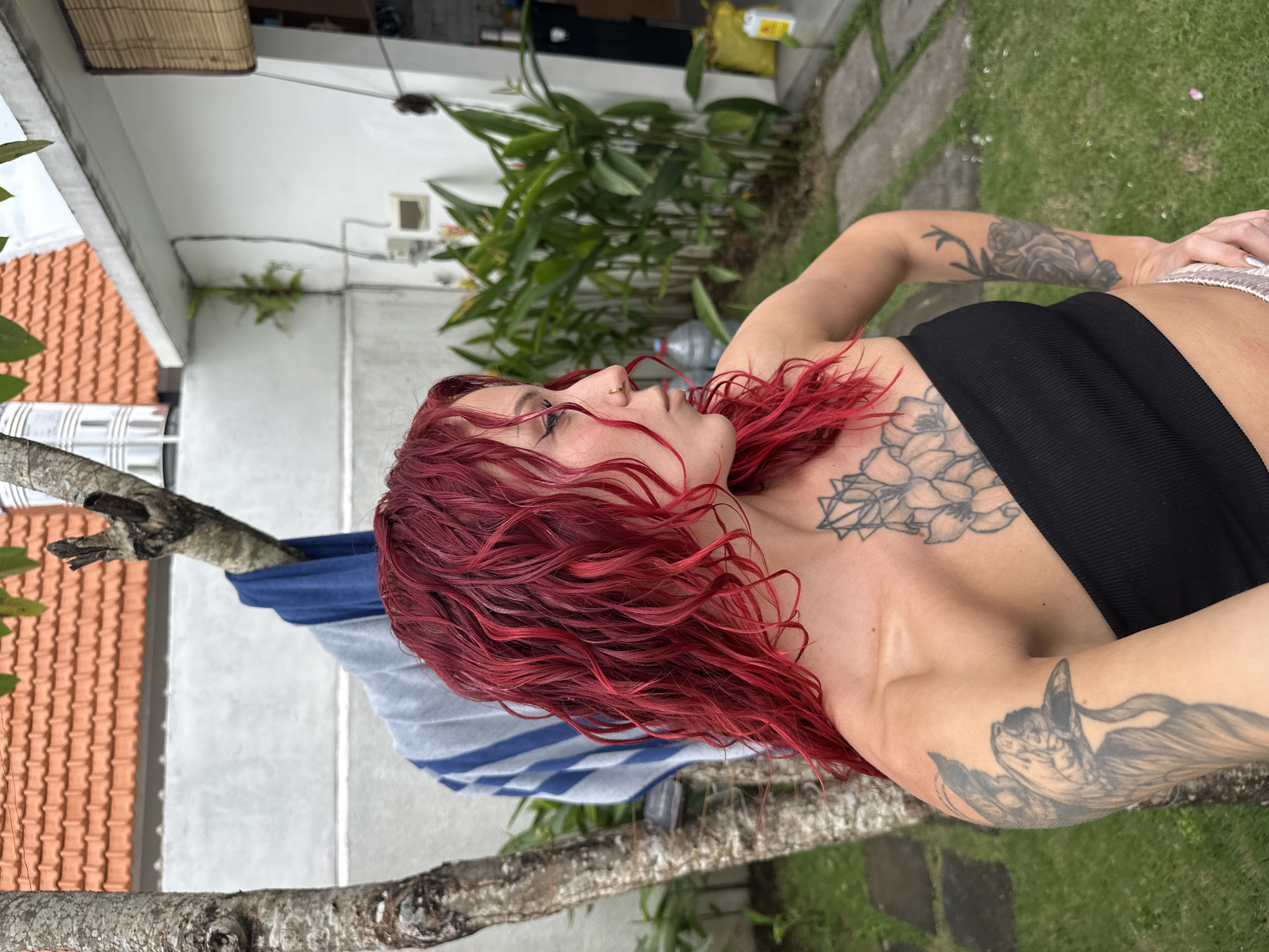 A young adult female is depicted in an outdoor setting in North Kuta, Indonesia, seen from the upper torso looking upwards and to her left. She has striking vibrant red, wavy hair that appears wet or freshly styled, and a nose ring. Multiple tattoos are visible on her body, including a floral design on her chest, a geometric pattern on her shoulder, and a stylized winged cat or animal head on her right arm. She is wearing a black ribbed bandeau-style top and light-colored bottoms.
The background reveals a residential scene with white-painted building walls and an orange tiled roof visible on the left. Green tropical foliage and plants are scattered throughout, with a grey stone path and patches of green grass underfoot. In the midground, a blue and white striped towel or garment is draped over a tree branch, suggesting laundry drying. Another tree branch frames the lower left foreground. The ambient lighting indicates it is daytime. The overall setting is a relaxed tropical backyard or courtyard. No visible text is present in the image.
