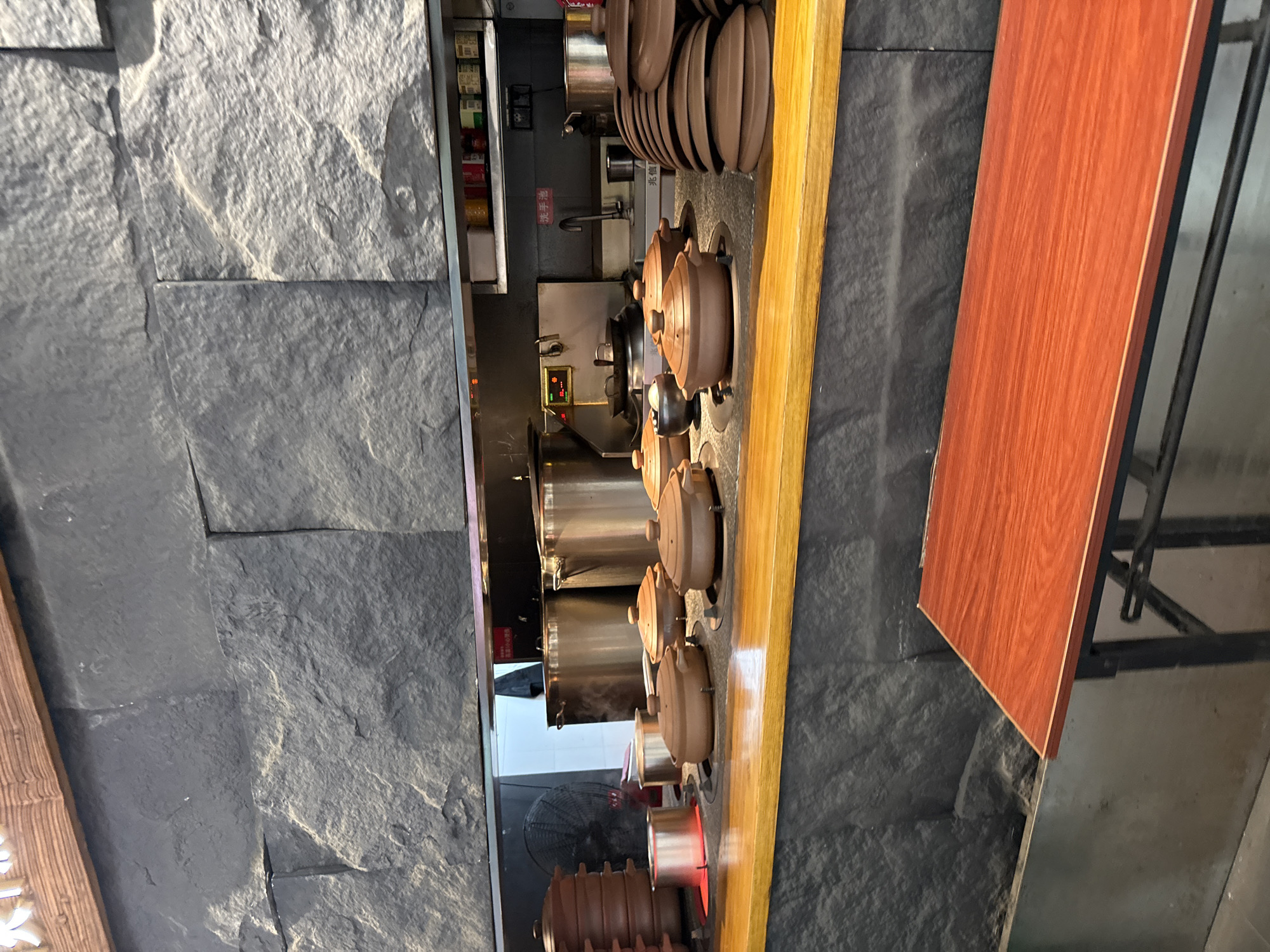 The image depicts a cooking station in what appears to be a restaurant, likely located in Tianhe District, China. The scene is dominated by a gray stone wall and a wooden countertop. On the countertop, several clay pots with lids are arranged, some of which are sitting on cooking elements that appear to be heated, indicated by a red glow underneath one of them. To the left, there is a stack of similar clay pots and some metal containers. In the background, large stainless steel pots are visible, along with a fan. Shelves above the cooking area hold various canned goods. A sink with a faucet is also present. In the foreground, a wooden table with a reddish-brown top and black metal legs is partially visible. There are no people in the frame. The overall impression is one of a functional kitchen space, possibly set up for self-service or a specific cooking style.