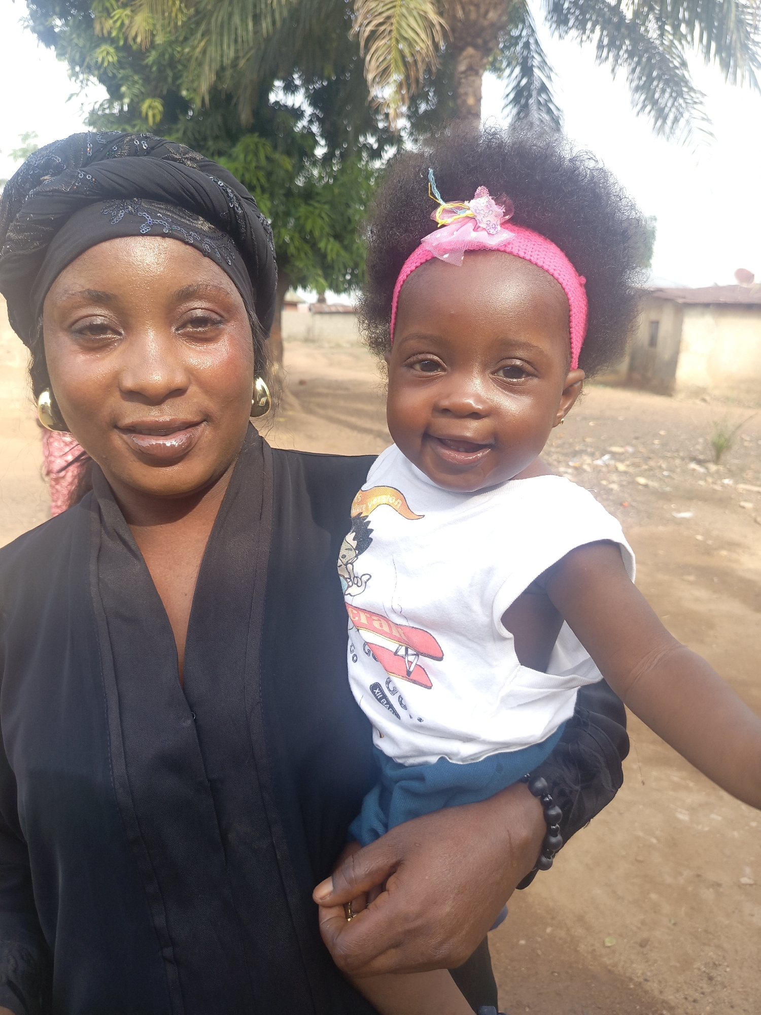 The image shows a woman holding a young child in an outdoor setting. The woman is wearing a black head covering and a dark-colored robe. She has gold hoop earrings. The child is wearing a pink headband with a decorative bow and a white t-shirt with a cartoon graphic. The background includes trees, a building, and what appears to be an unpaved ground surface. The specific location in Akwanga, Nigeria, cannot be determined from the image. The scene suggests a casual, daytime moment.