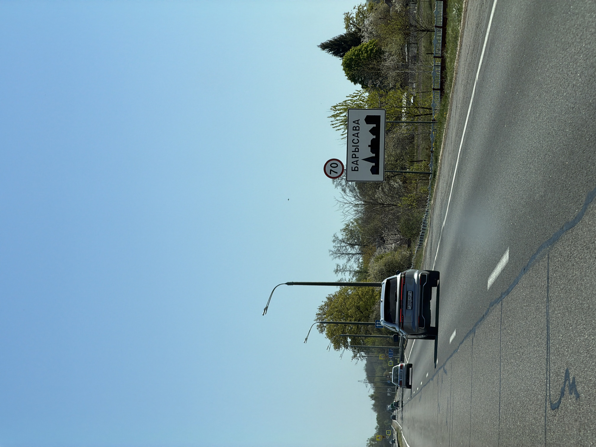 The image depicts a highway in Кіселявецкі сельскі Савет, Belarus, approaching the city of Barysaŭ. A sign indicating the city's name in Cyrillic (БАРЫСАВА) and a speed limit of 70 km/h is visible on the roadside. Cars are traveling on the multi-lane road, with a gray SUV directly in front of the camera. Overhead streetlights are present along the roadway. The sky is blue, suggesting clear weather conditions. Trees and vegetation line the side of the road beyond the sign.