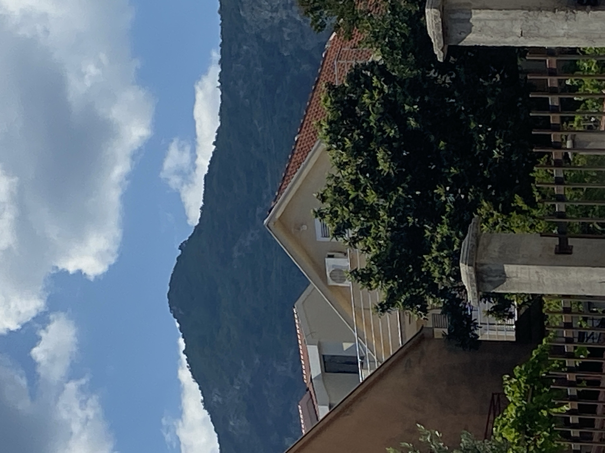 This image shows a view of residential buildings in Budva, Montenegro, with a large mountain in the background under a cloudy blue sky. The foreground features the tops of houses with terracotta roofs, balconies, and green trees. A dark metal fence and a concrete pillar are also visible in the lower part of the frame. The overall impression is one of a sunny day in a coastal town with mountainous terrain.