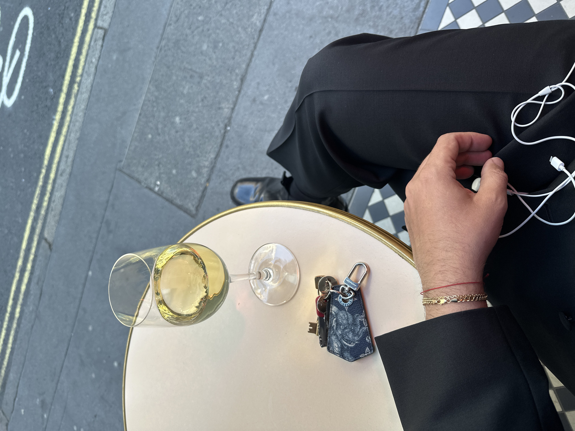 An overhead, slightly angled shot captures a person seated at an outdoor cafe table in London, United Kingdom, viewed from a first-person perspective. The person is wearing black trousers and a black jacket, with black shoes visible beneath the table.
On the small, round table with a light top and golden rim, a glass filled with white wine is prominently placed. Beside it lies a set of keys attached to a distinctive keychain fob. The fob is dark with a patterned design, and a silver clasp clearly displays the text "LOUIS VUITTON". The person's right forearm and hand are visible, adorned with a thin red string bracelet and a gold chain bracelet. White wired earphones are draped across the person's lap.
The outdoor setting includes a street or pavement area on the left, marked with double yellow lines characteristic of UK roads. Some white, illegible graffiti is visible on the asphalt. Beneath the table and extending to the right, a black and white checkerboard tiled floor is visible, suggesting a cafe or restaurant entrance. The scene is illuminated by natural daylight, indicating clear weather. The overall impression is one of leisure, with the person enjoying a drink outdoors.