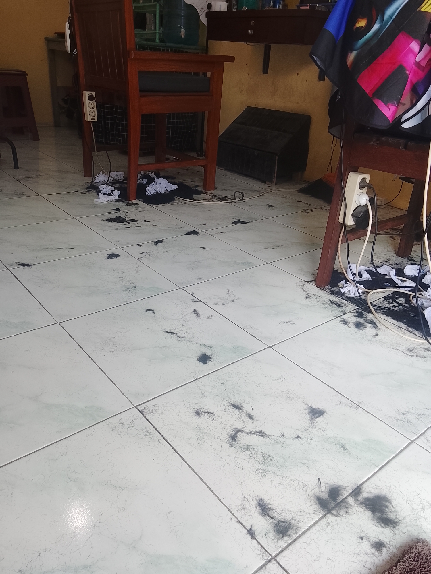 The image depicts the floor of what appears to be a barbershop in Karangklesem, Indonesia. The floor is tiled and covered in scattered hair clippings, indicating recent activity. Two wooden chairs are visible, one in the foreground and another further back. Electrical outlets with cords are attached to the chairs, suggesting they are for grooming equipment. A colorful cape hangs over the chair in the right foreground. A dark, folded object, possibly a hair trap or a barber's station, is situated on the floor near the back wall. The overall scene is one of a working barbershop, albeit one that requires cleaning.