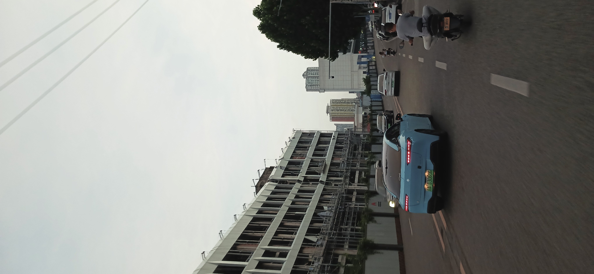 The image shows a street scene in Jiulongpo District, Chongqing, China. A light blue car is traveling on a paved road, with several buildings visible in the background, including a tall unfinished structure to the left. A motorcycle and other vehicles are also on the road. The sky is overcast.