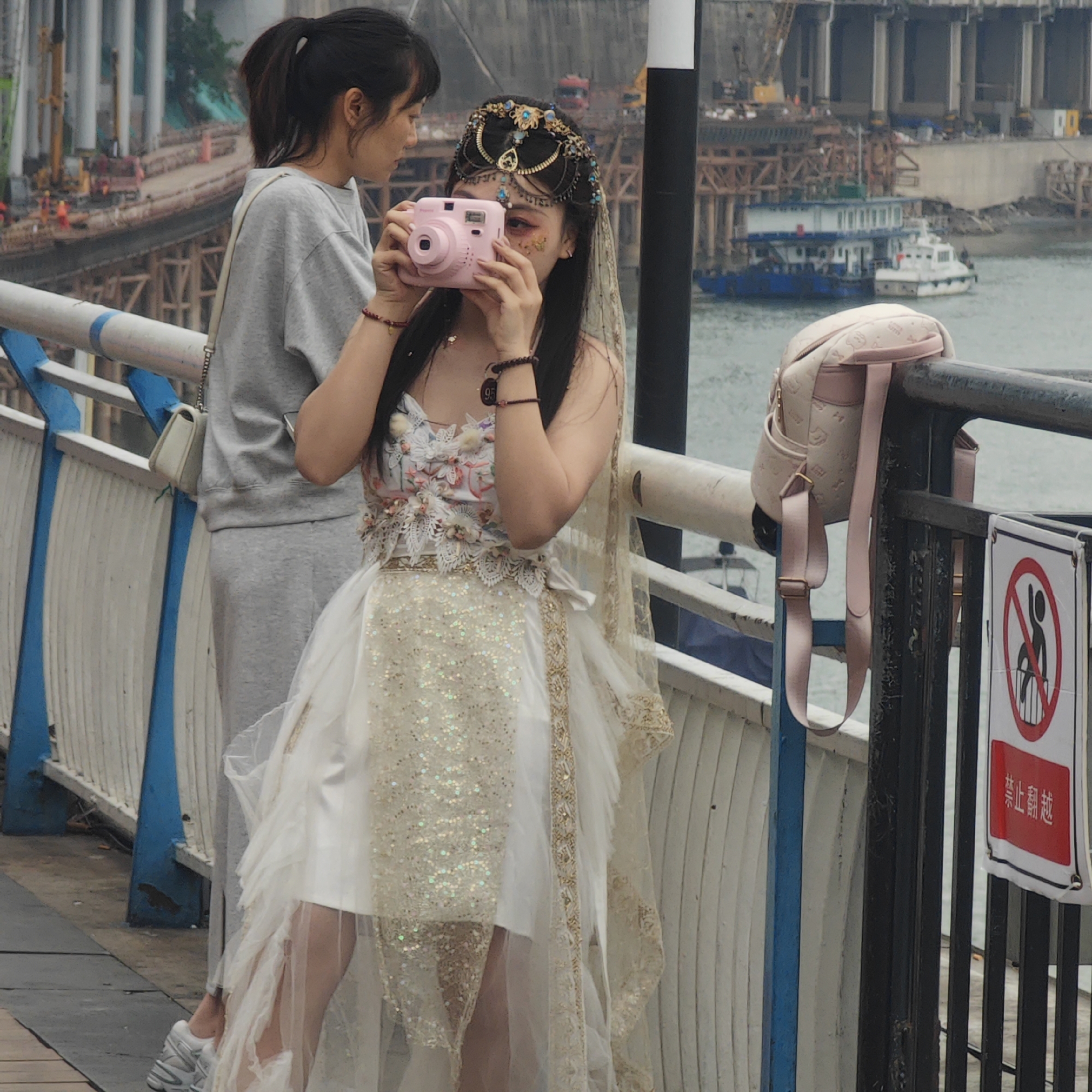 On a bridge in Yuzhong District, China, two women stand near a railing, overlooking a body of water with boats and waterfront construction in the background. The woman in the foreground is dressed in an elaborate, ornate white and gold dress with floral embellishments and a decorative headpiece, holding a pink instant camera up to her face as if taking a photograph. The woman behind her is dressed in a casual grey tracksuit. A light pink backpack hangs on the railing near a sign that warns against climbing over.