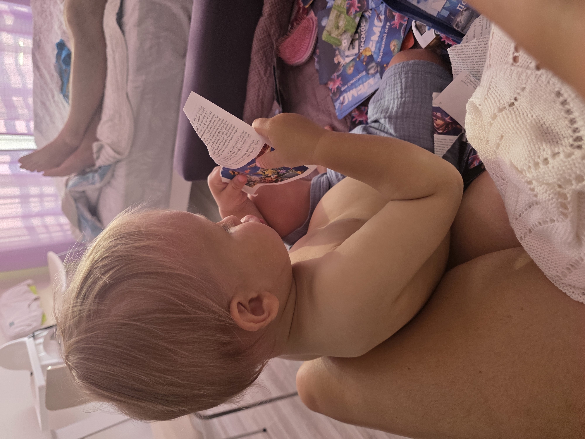 The image shows a close-up view of a young child, likely a toddler, held by an adult. The child is sitting and looking at a colorful booklet or magazine. The booklet appears to be about marine life, with images of fish and coral visible. The child's attention is focused on the booklet, and their expression suggests curiosity or engagement.
The setting appears to be indoors, possibly a home or a hotel room, given the presence of soft furnishings like blankets and a bed. The lighting suggests it could be daytime, though no specific weather conditions are discernible. A white baby chair or stand is partially visible in the background.
The adult's arm and leg are visible, supporting the child. A white knitted garment, possibly a shawl or blanket, is draped over the adult. The overall atmosphere is intimate and suggests a moment of quiet play or learning.
Visible text on the booklet is in Russian and mentions "Подводный мир" (Underwater world) and "КОСМОС" (Cosmos), suggesting it's an educational or entertainment publication for children. There are also snippets of text related to marine animals, coral reefs, and scientific facts about the ocean. The child's hair is blonde and fine. The scene conveys a sense of tenderness and focused attention on the learning material.