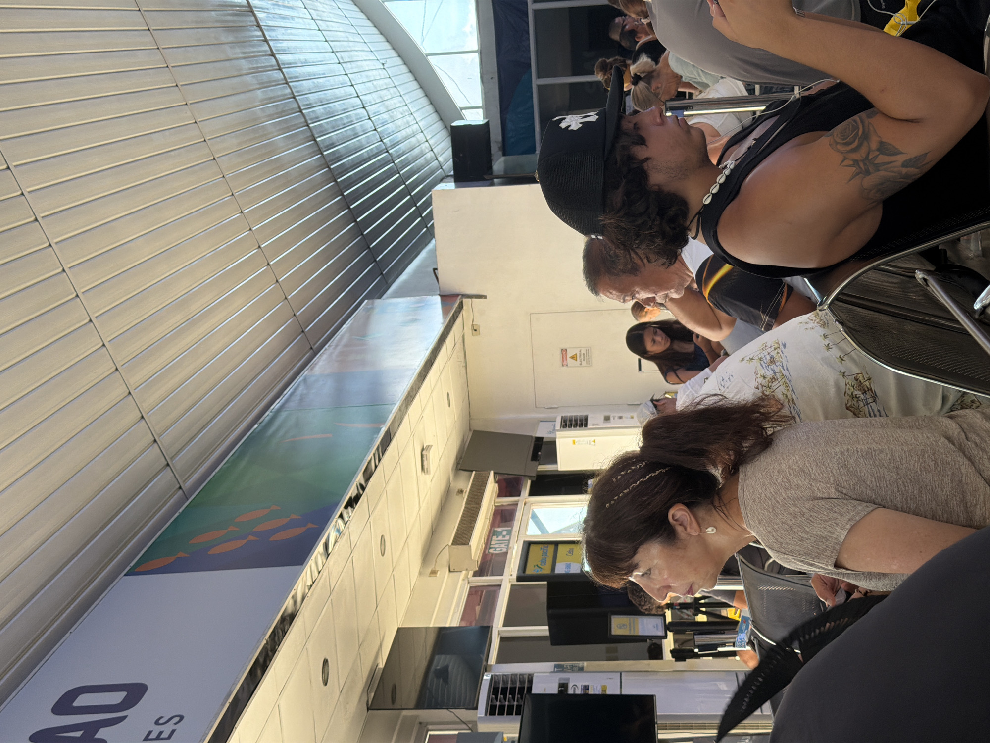 The image shows a crowded waiting area, potentially at an airport or bus terminal, in Del Carmen, Philippines. Multiple people are seated and standing. A man in the foreground wears a black tank top and a backward black baseball cap, showing a tattoo on his upper arm. Behind him, a diverse group of people occupies the available seating. The environment includes a wall with horizontal panels, possibly part of a gate designation, and information displays. Signs indicating "GATE-..." are visible. The image appears to have been taken from a low angle and rotated, making it difficult to ascertain the exact layout of the space.