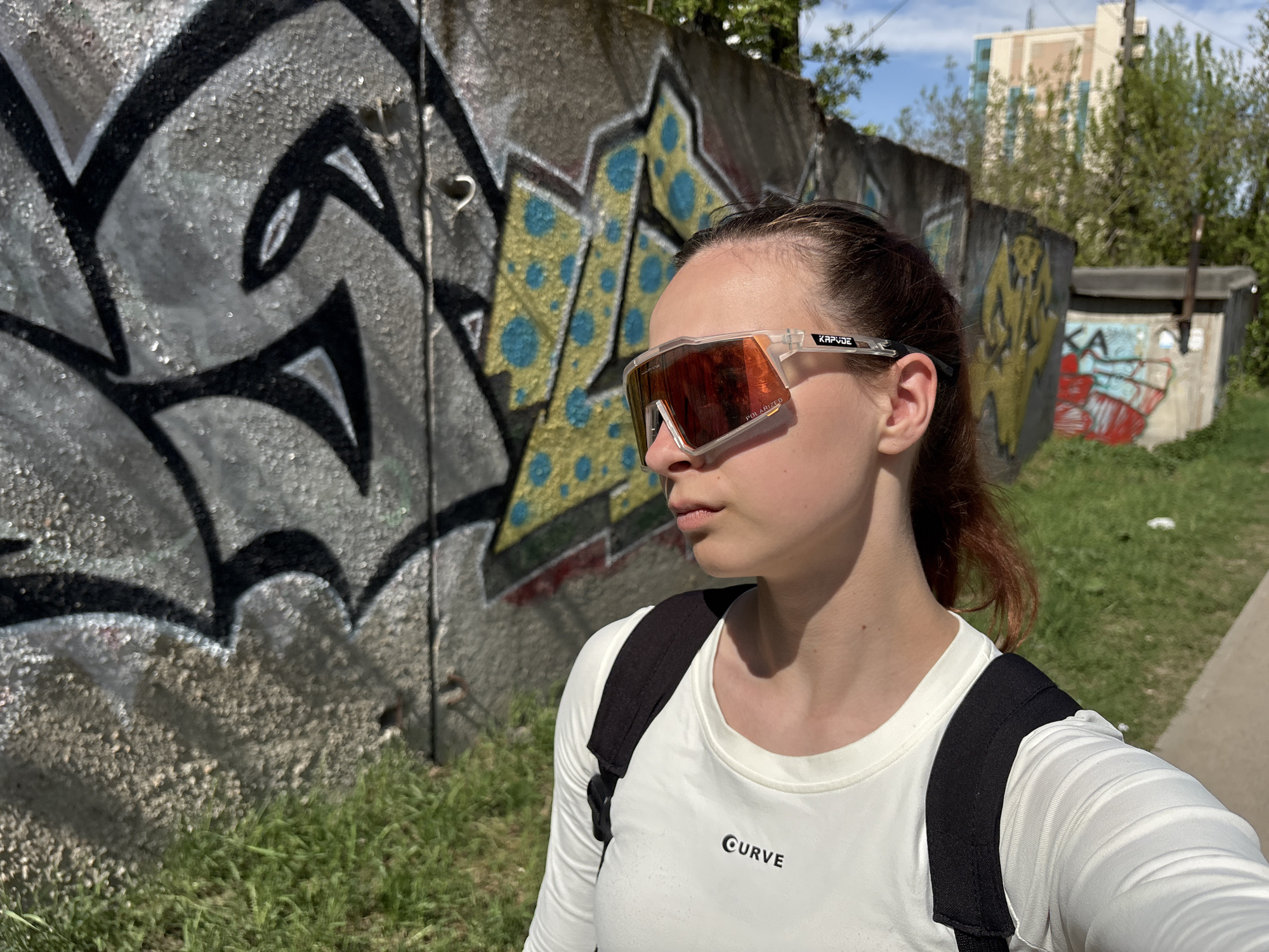 A medium shot depicts a fair-skinned woman wearing sunglasses, a white long-sleeved shirt with the word "CURVE" printed on it, and a black backpack. She stands on a narrow sidewalk, facing slightly away from the camera towards the left, against a concrete wall covered in graffiti. Patches of grass grow along the base of the wall and sidewalk. In the background, beyond the wall, a small outbuilding with more graffiti and trees are visible, with a taller building in the distance. The lighting suggests a bright day. Based on the graffiti style and architectural features, the image is likely taken in Perm, Russia.