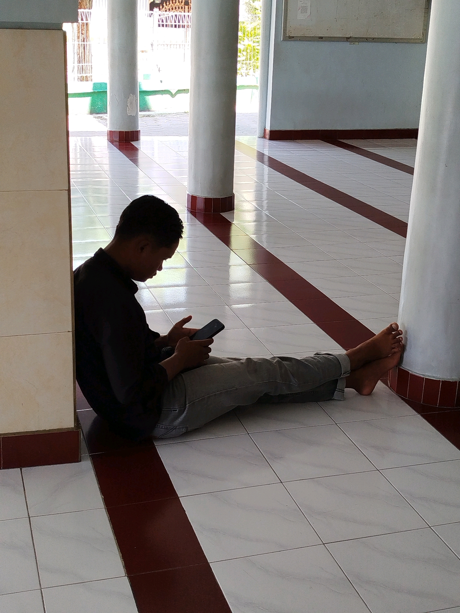 A young man, dressed in a black long-sleeved shirt and grey jeans, sits on the tiled floor of an open-air building, his back leaning against a cream-colored column. He is looking down at his mobile phone, which he holds in his right hand. His legs are extended out in front of him, and his bare feet are resting against another large white column. The floor is tiled with white squares and bordered by maroon stripes. Sunlight streams in from an open doorway in the background, casting long shadows across the floor. The environment appears to be a public space, possibly a mosque courtyard or a waiting area in Surabaya, Indonesia, given the architectural style and the presence of similar pillars and tiled flooring often found in such locations.